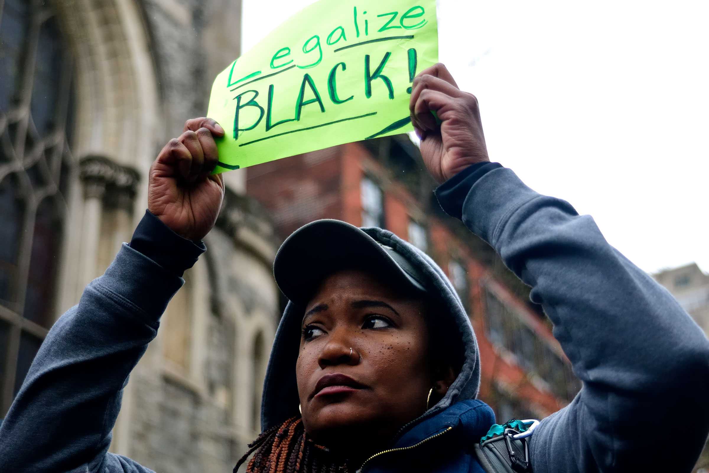 A woman looking defeated and sad is holding up a small neon green sign that says "Legalize Black".