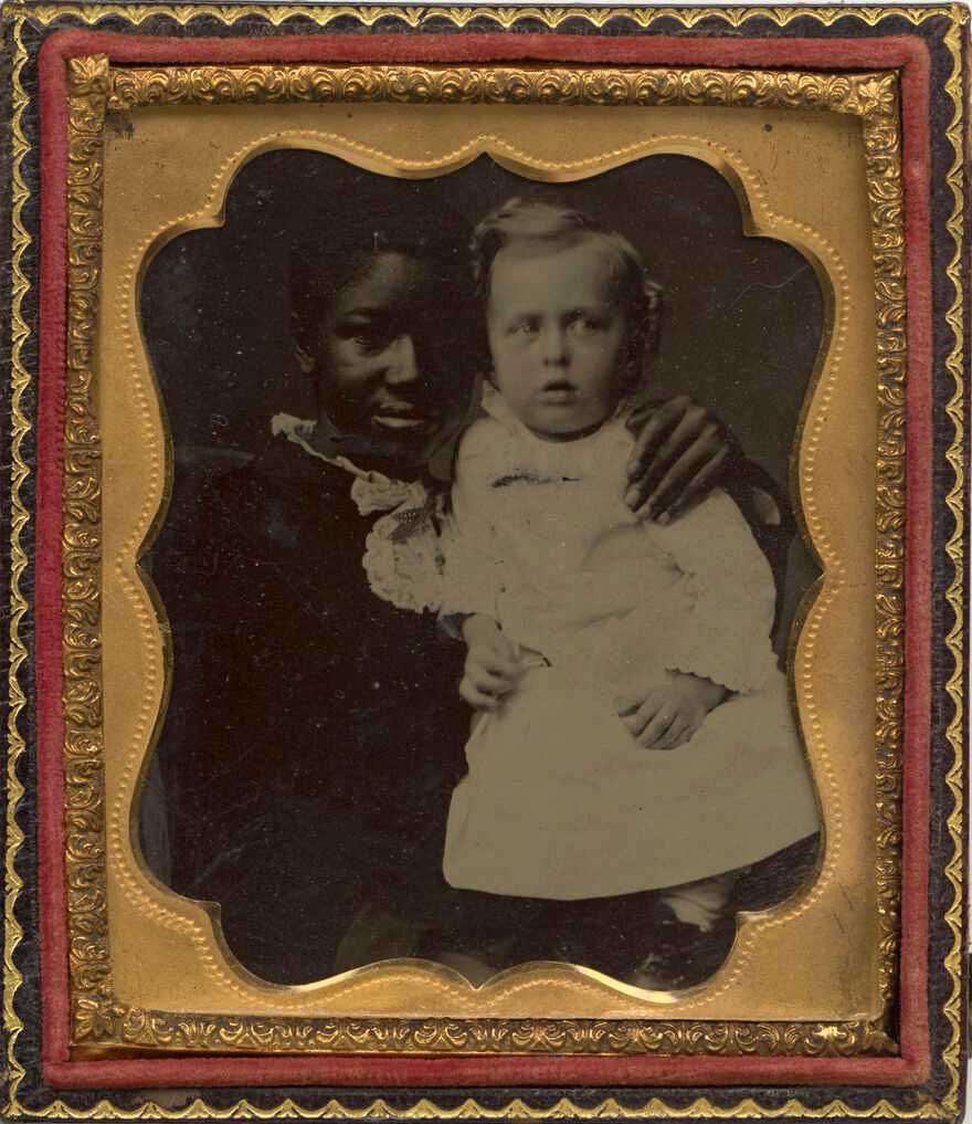 This 1/6 plate tintype photograph depicts a young black woman with a white baby. The young woman is seated, wearing a dark colored dress and light colored lace collar. She stands the baby up on her lap. The baby wears a long, light-colored dress and has tinted pink cheeks. The tintype is housed in a wooden case with gilt highlighting, gold colored metal framing and red velvet lining.