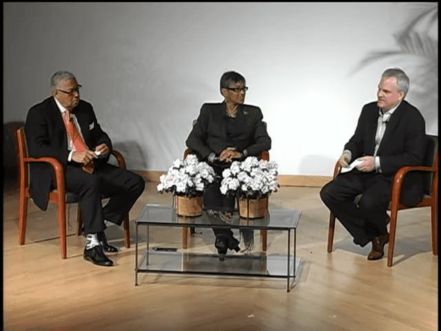 Screenshot from video showing two men and one woman seated before a glass coffee table with flowers on top.