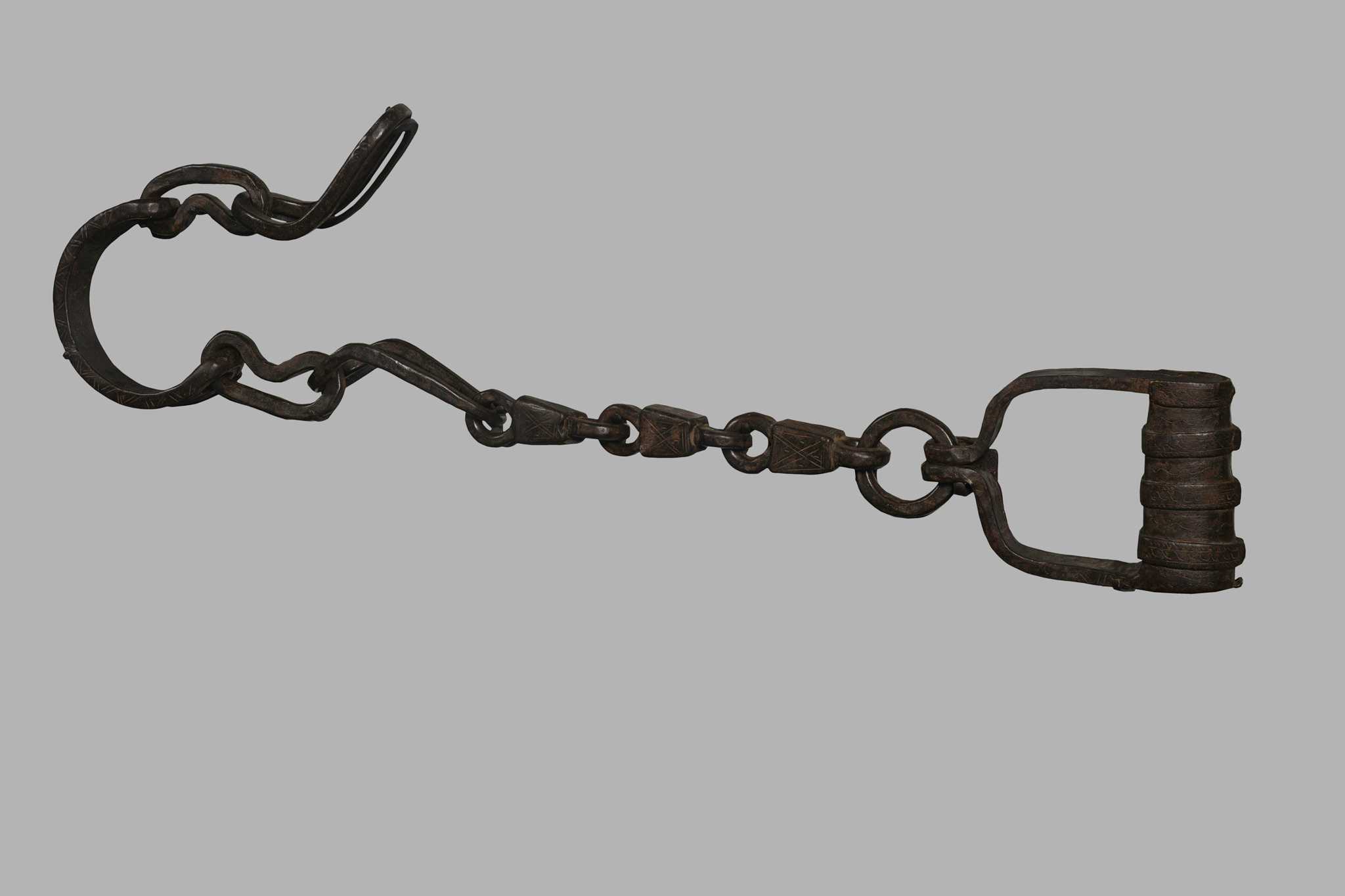 These ankle shackles consist of a two-piece cuff-like section at one end, and another cuff-like piece and locking device at the other; connected by three very heavy and elaborately incised links; all portions of this pair of shackles bear designs that are a series of parallel lines, some crossed into X-shapes.