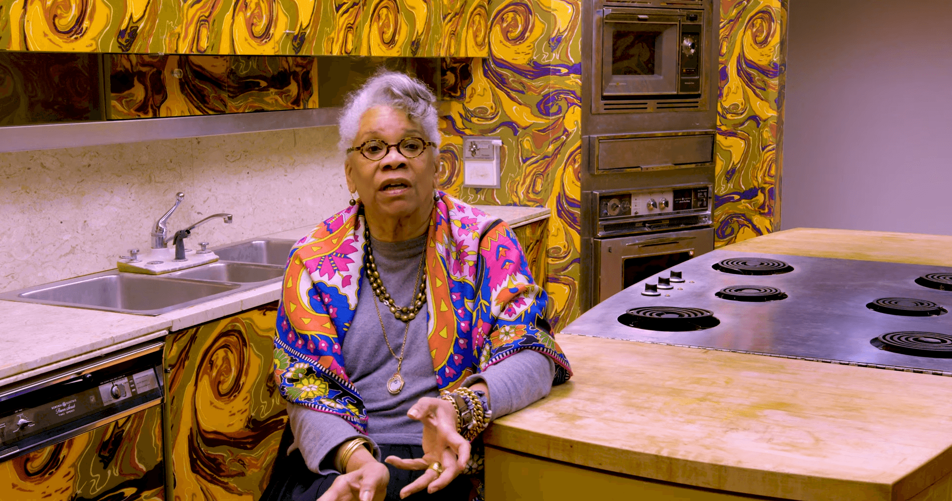 Dr. Jessica Harris is sitting in the test kitchen's counter with the colorful cabinets and countertop behind her.
