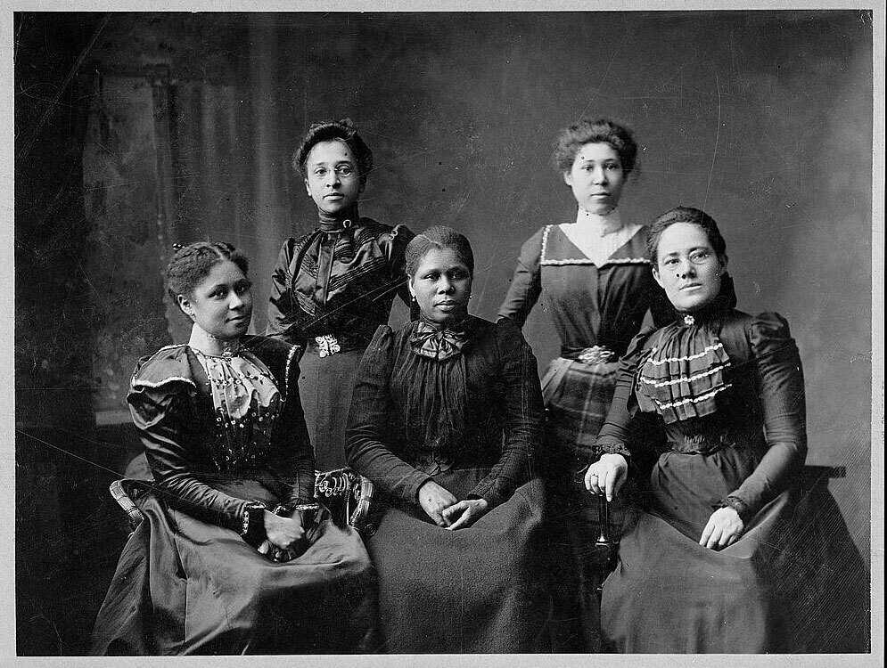 Black and white group portrait of 5 African American women in stylish dresses.  Three women are seated in front of two women standing behind them