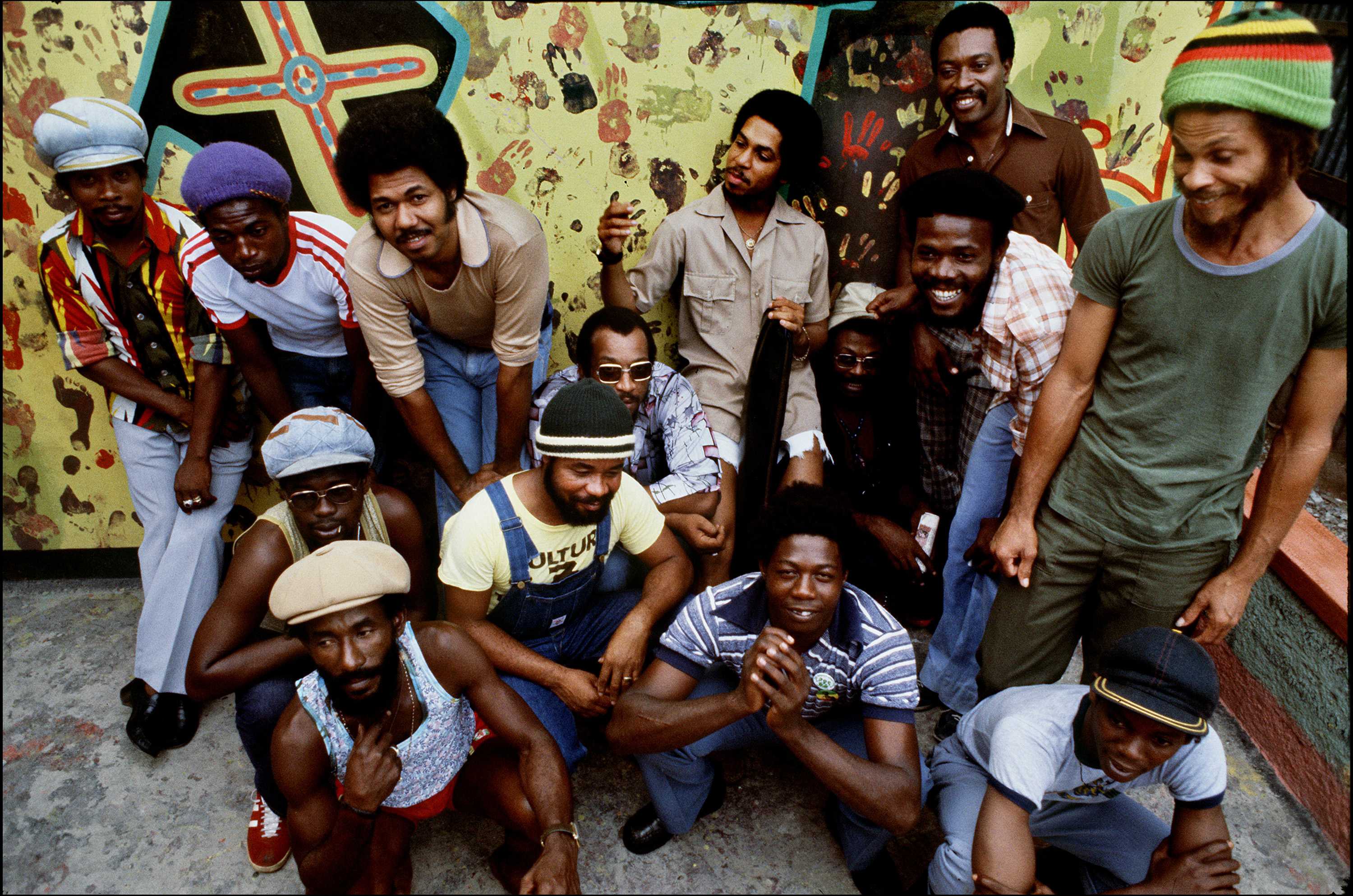 Lee “Scratch” Perry standing with members of The Upsetters outside. Some are crouching, others are standing in front of the yellow wall.