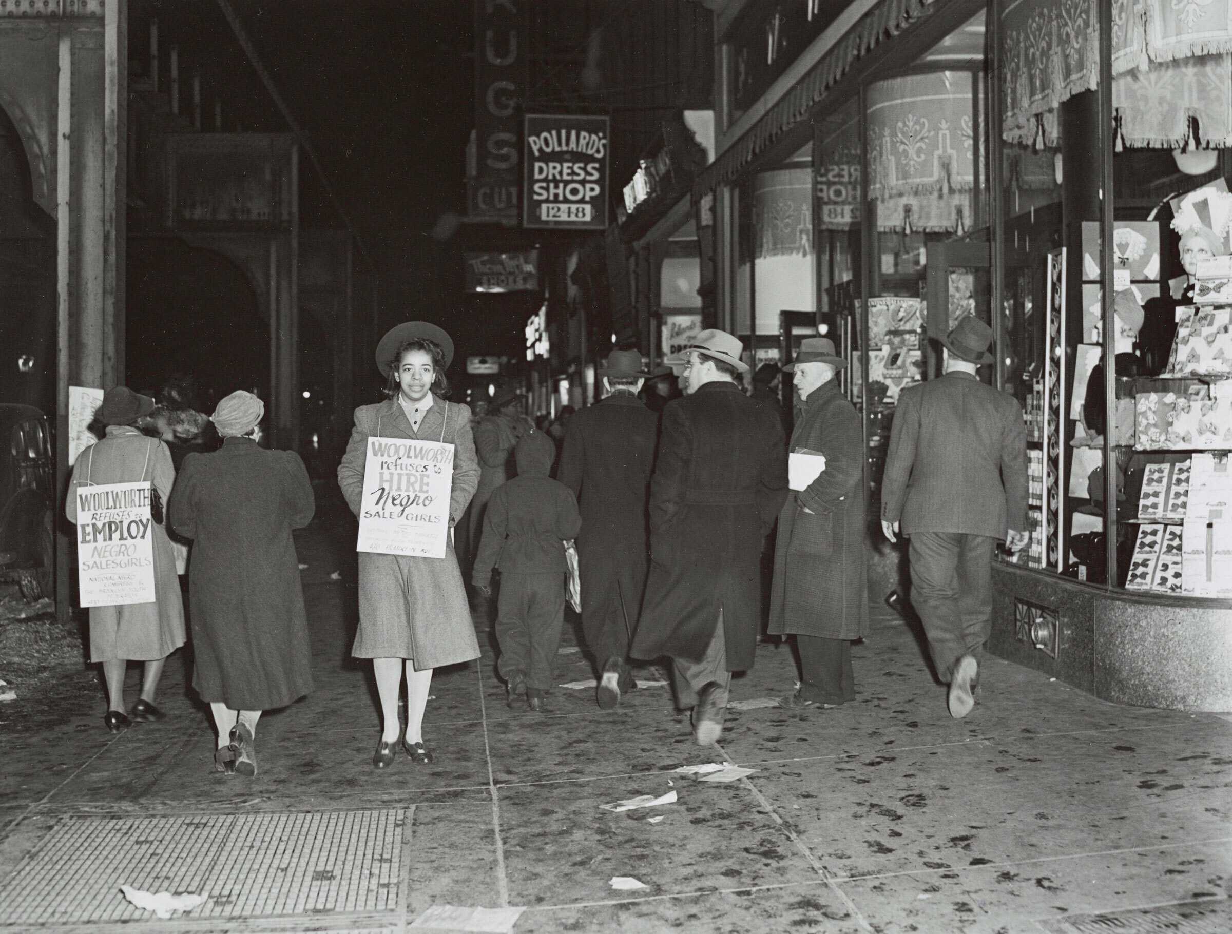A black-and-white photograph of two girls protesting in front of an F. W. Woolworth's store. The pair wear signs announcing that "Woolworth refuses to hire Negro sale girls."