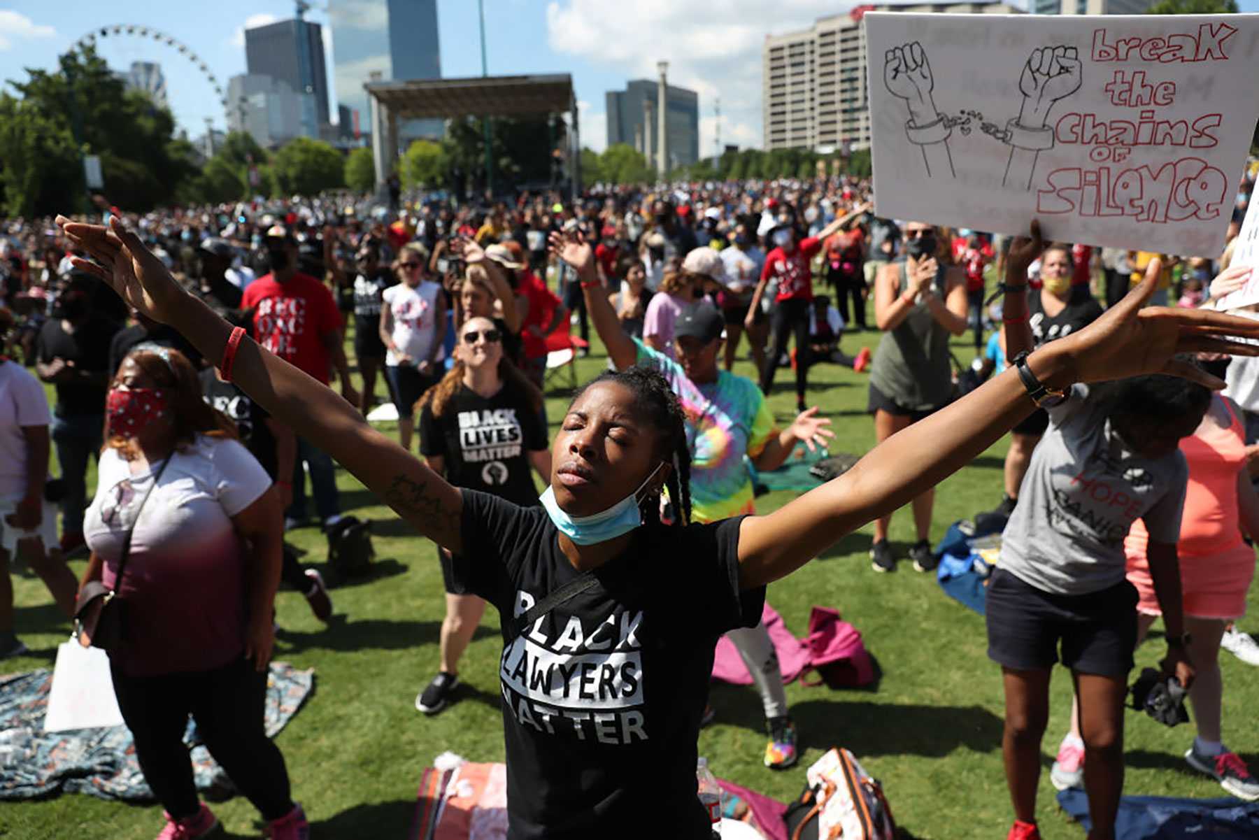 Crowd gathered in a park for a protest; person in foreground with arms raised, wearing a "Black Lawyers Matter" shirt.