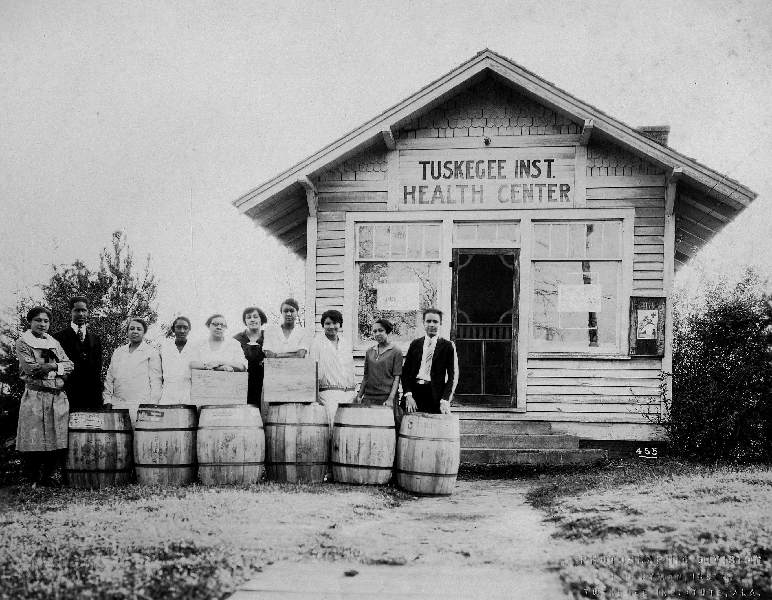 Photgraph of people standing in front of Tuskegee Institute Health Center with large barrels, circa early 20th century.