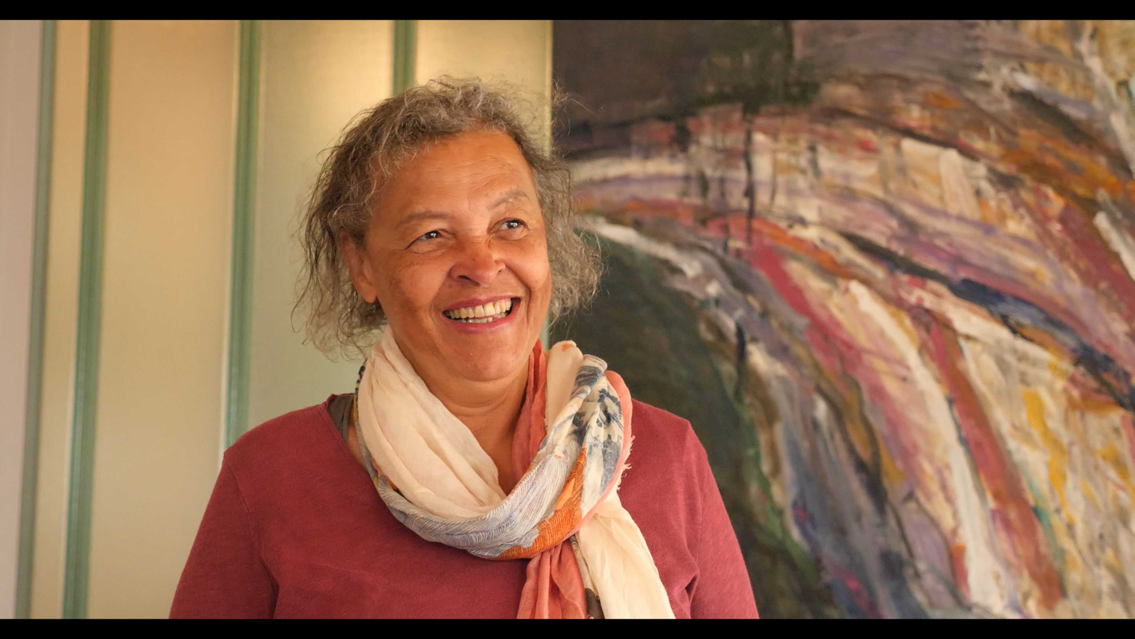 Person wearing a scarf standing indoors in front of a large colorful abstract painting.