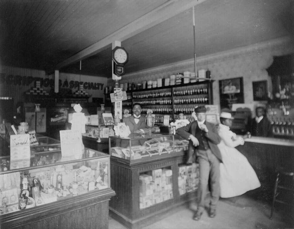 Interior view of drug store with African American male employee wearing bow tie standing behind the counter.  There are several other people in the photo including a man standing in front of the counter and a woman in a hat and long white dress at soda fountain.