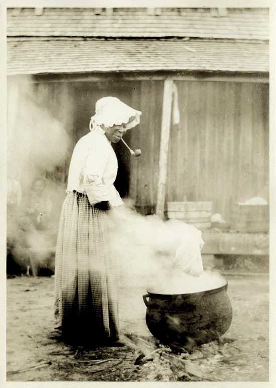 A sepia toned photo on a older woman using a hot cauldron. She has a pipe in her hand.