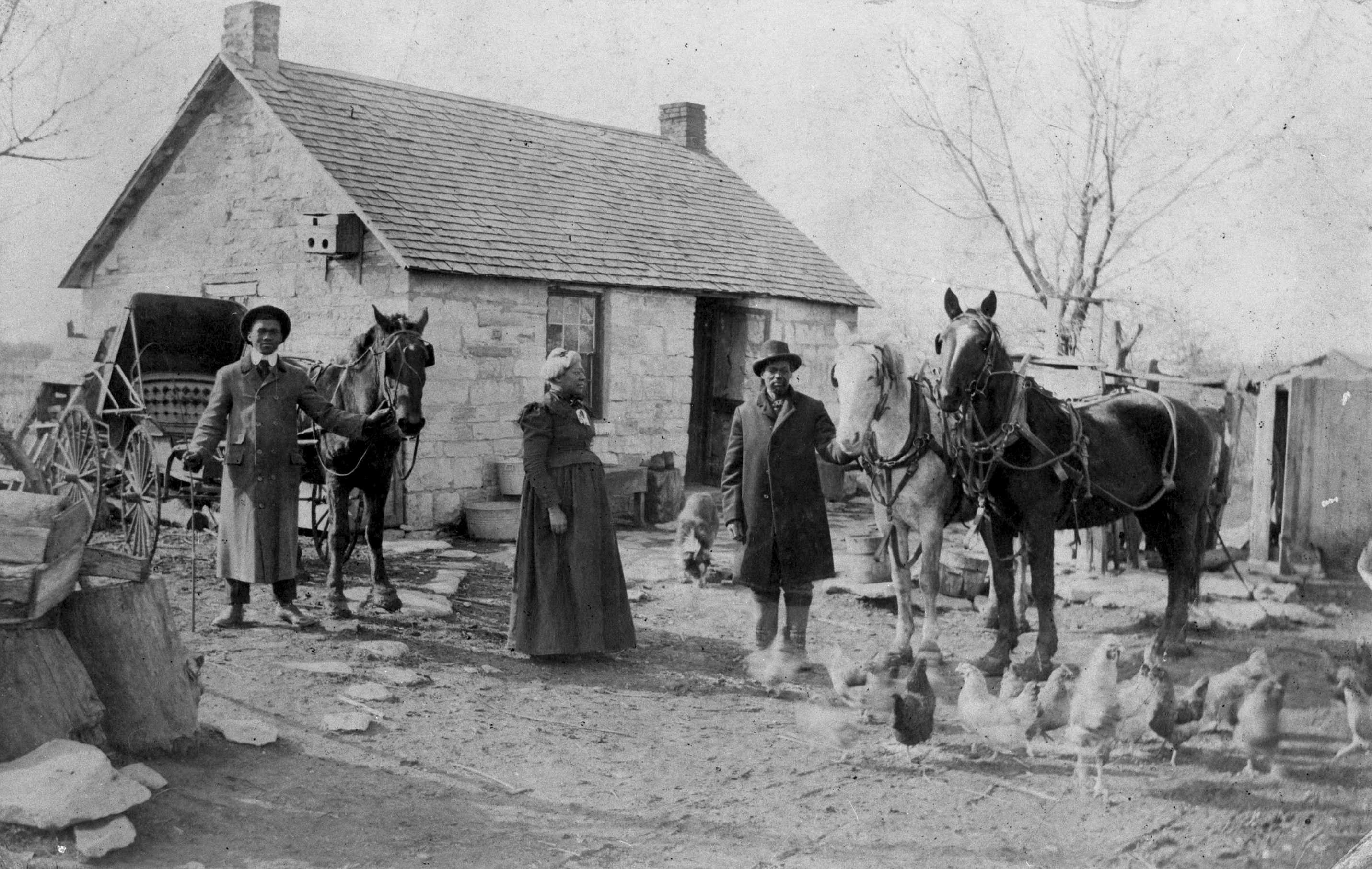 A black and white photo of two men hold horses.They are surrounded by chickens and other farm items.