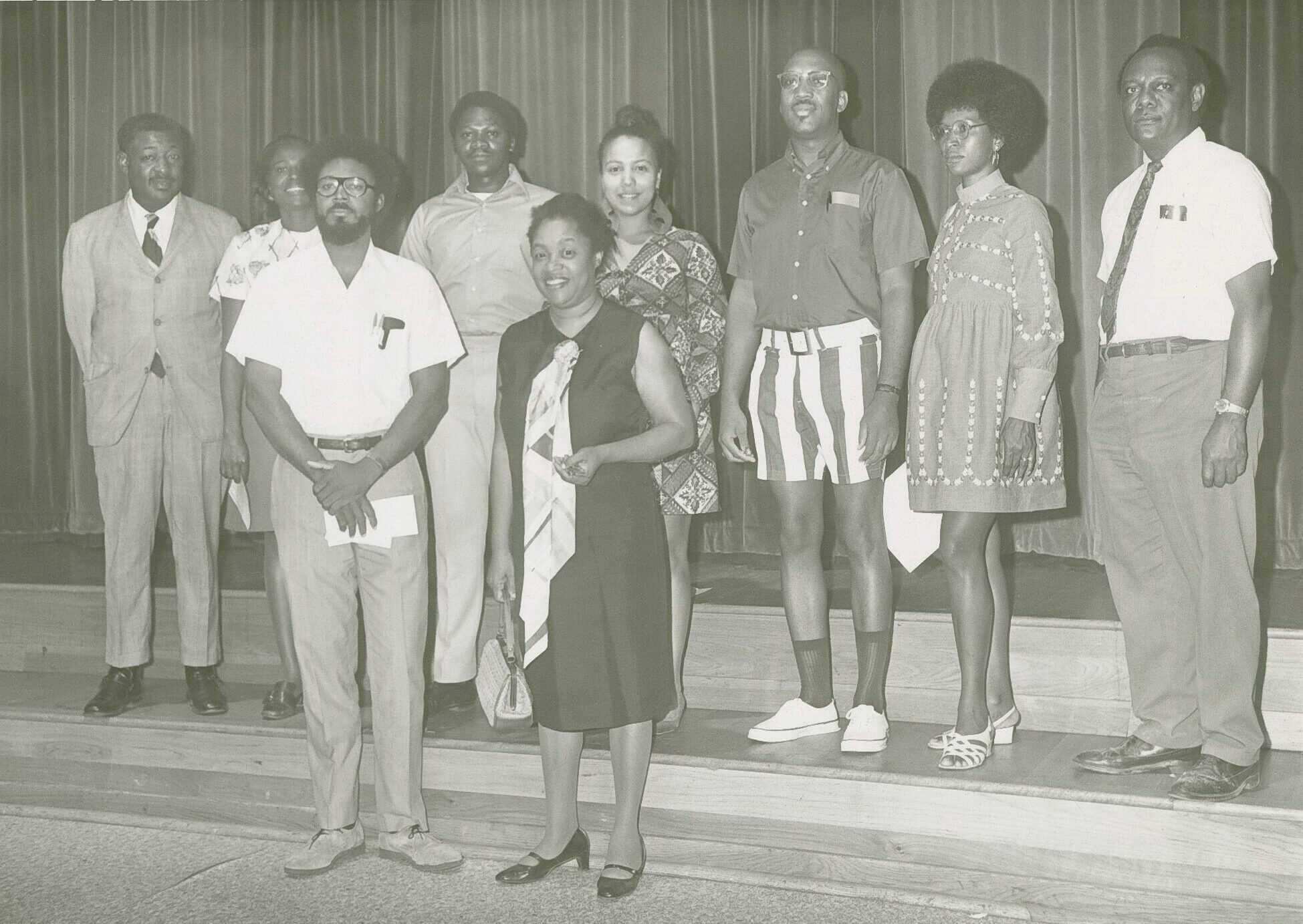 Eight individuals standing on a stage in front of a curtain, arranged in two rows—five in the back and three in the front—wearing a mix of formal and casual attire, suggesting a group event or presentation in an indoor auditorium.