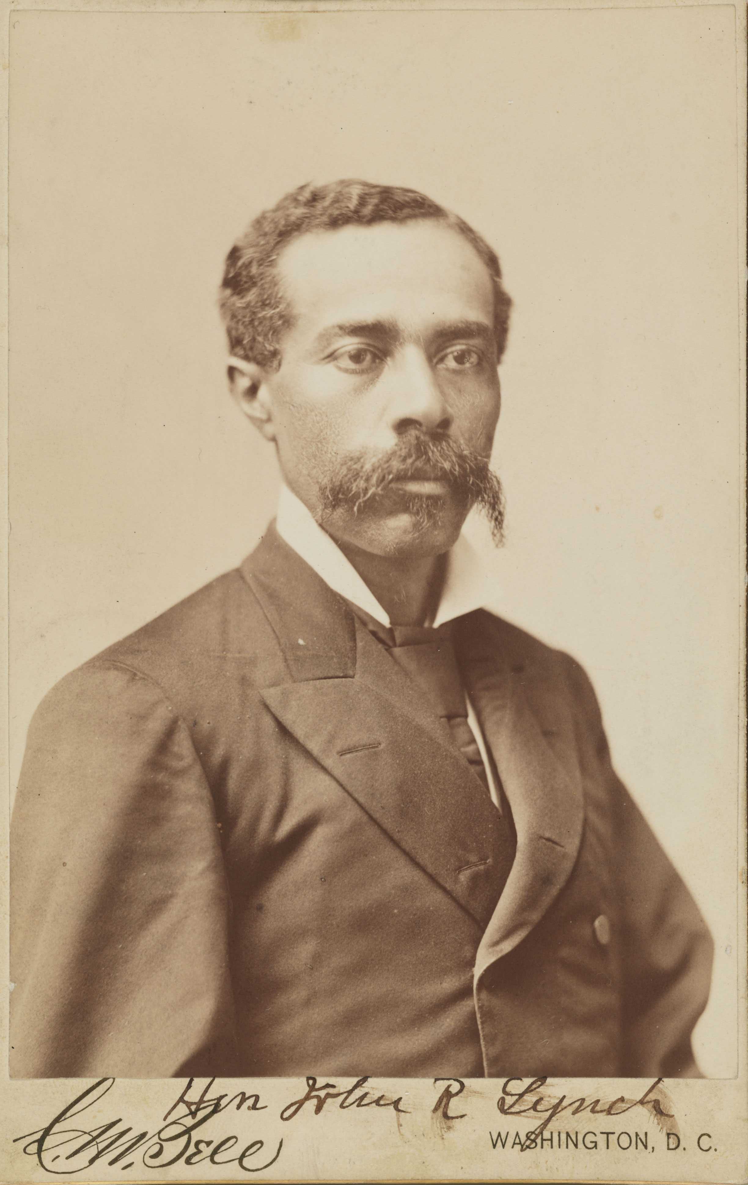 Sepia toned, half length portrait of African American man with large hand bar mustache.  Below the image "Hon John R Lynch" is handwritten and "Washington, D.C." is printed.  The image is also signed by the photographer "C. M. Bell"