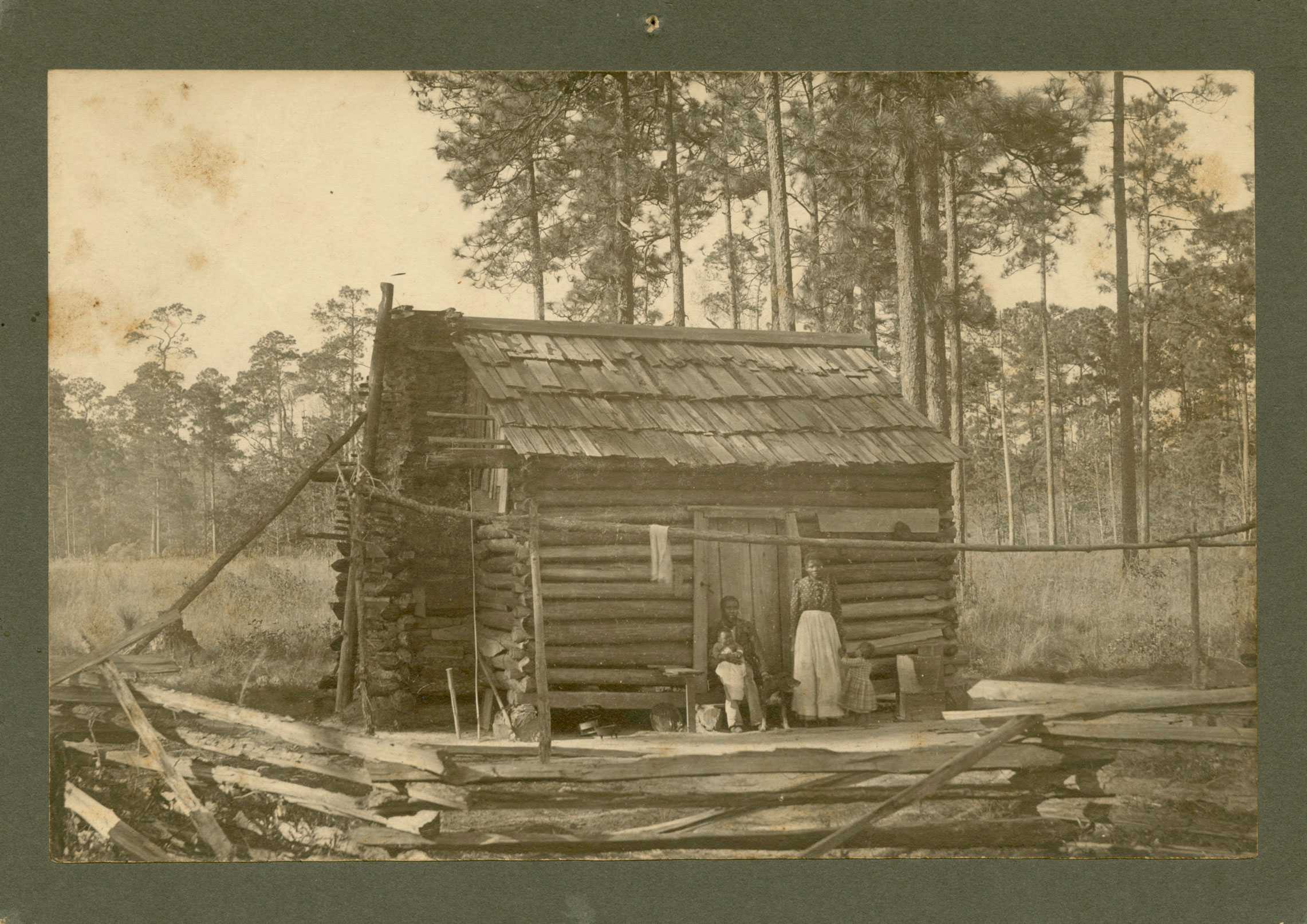 Black and White photograph of a family in front of a log cabin with dog.