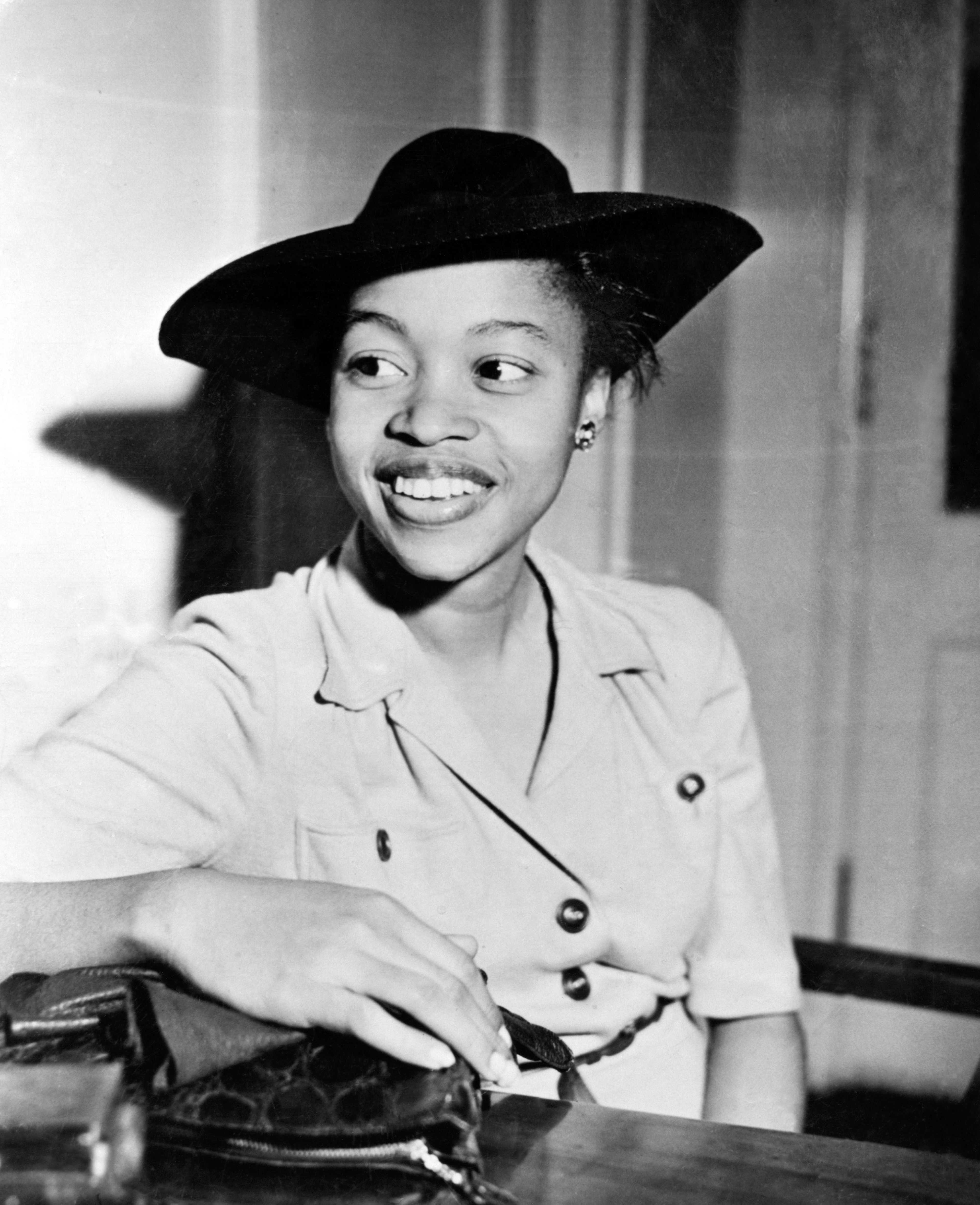 Author Margaret Walker seated at a table wearing a light-colored buttoned dress and a dark wide-brimmed hat, looking to the side with an open-mouthed expression.