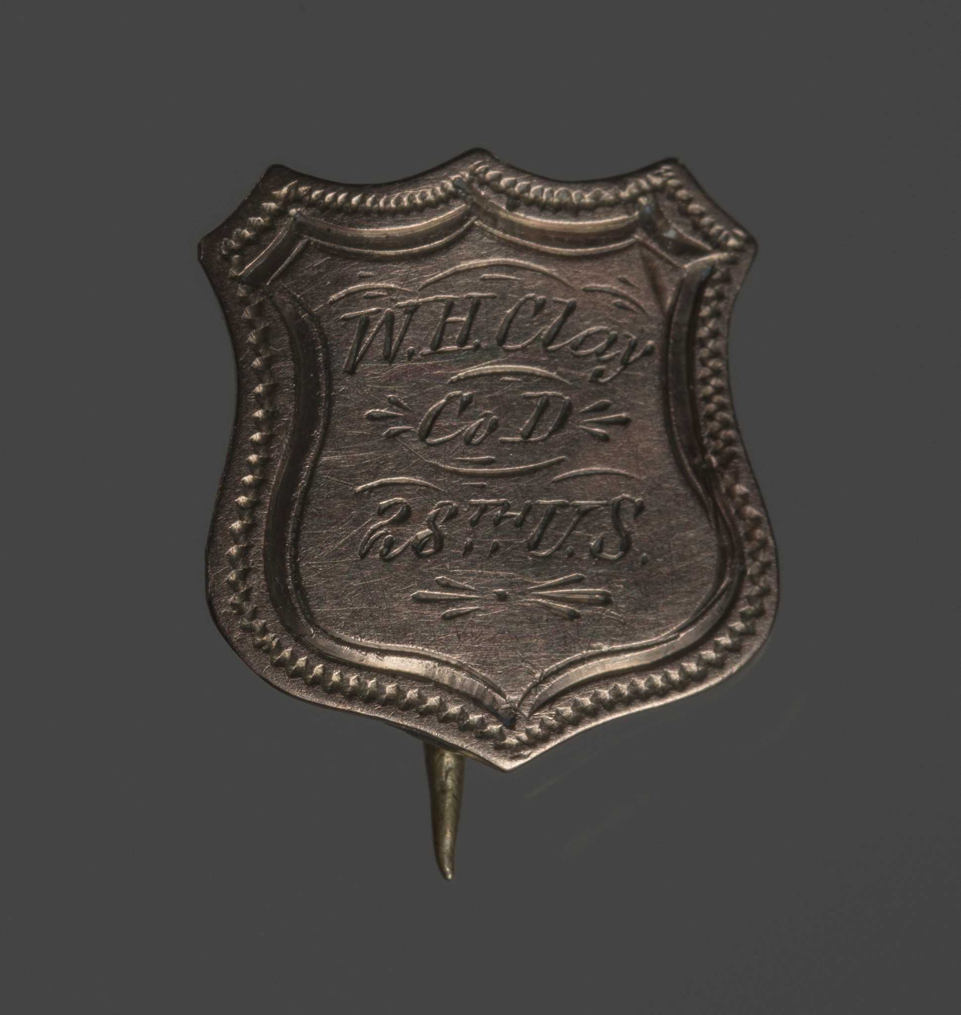 A shield-shaped silver metal badge with stick-pin back. The badge decorative borders around the edge and is engraved in the center: "W. H. Clay Co. D 28th US."