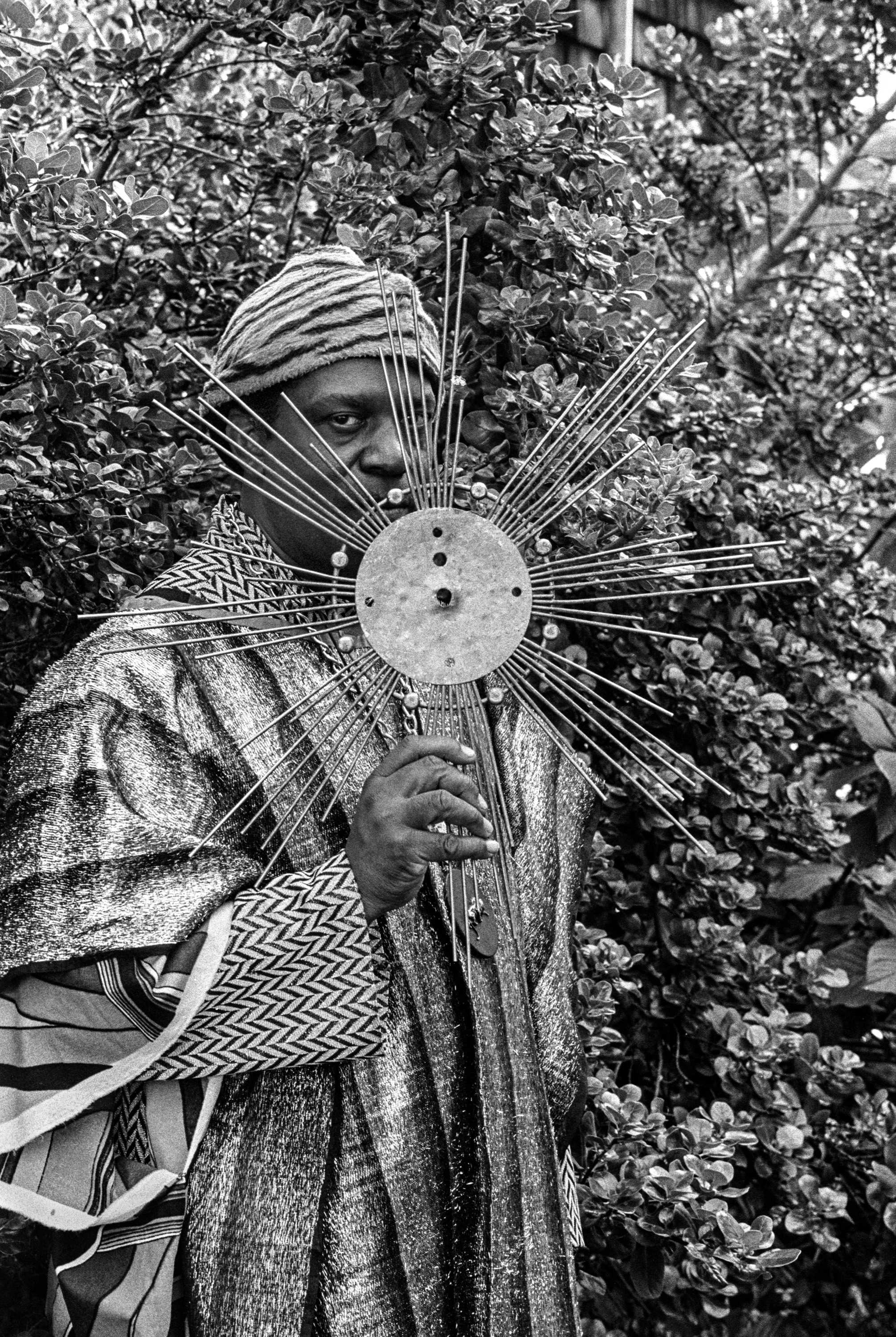 Sun Ra posing for a black and white photo with a large metal structure that looks like a sunburst in front of his face.