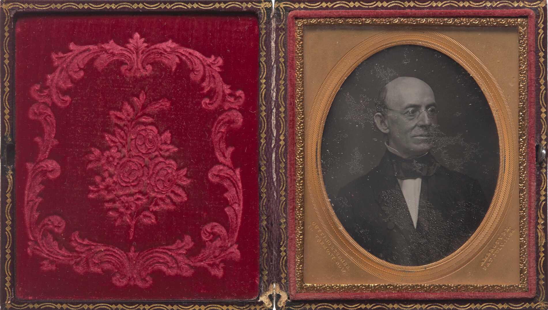 A daguerreotype of William Lloyd Garrison in a case. In the portrait, Garrison is wearing a suit and glasses and looking off to the left. The exterior of the case is black with gold trim. The interior has embossed red velvet on the left side and the photograph inside a gold oval frame on the right. The glass over the image is heavily scratched. Stamped into the bottom left corner of the frame: "CUTTING & TURNER / 10 TREMONT ROW". And in the bottom right corner: "AMBROTYPE. / PAT. JULY. 11-54"
