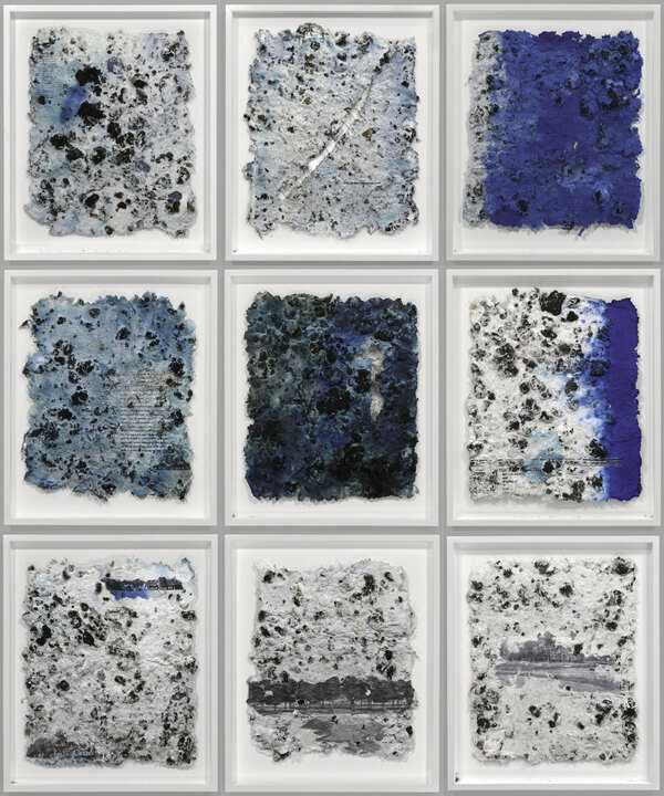 Nine framed abstract artworks arranged in a 3×3 grid, each showing textured, irregular square panels in white, gray, black, and deep blue with rough, speckled surfaces and layered paint or material effects.