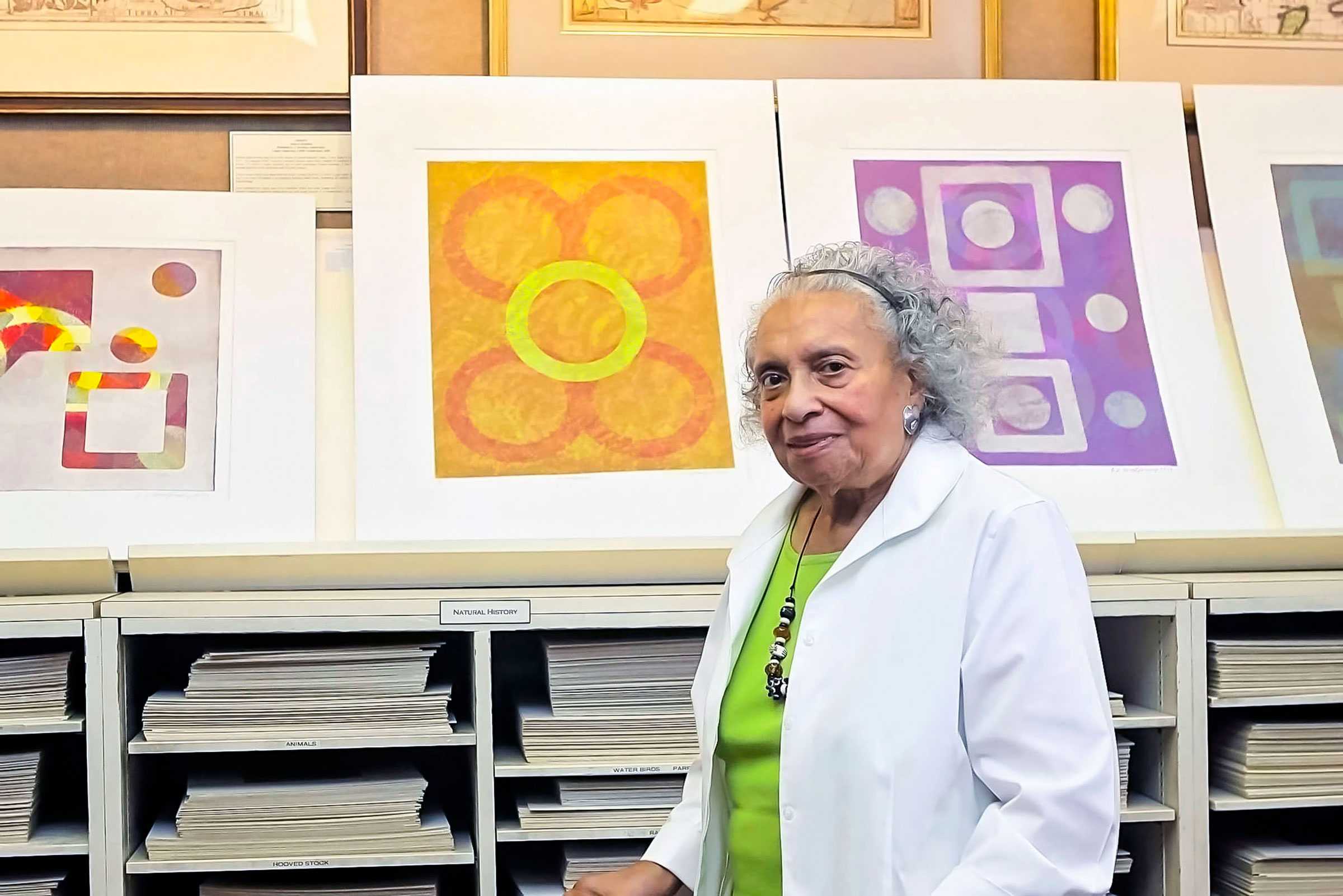 An artist stands beside abstract colorful artworks displayed on a shelf in a studio or gallery setting.