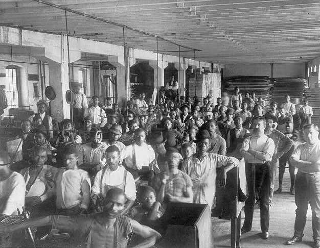 Black and white photograph of mostly African American males standing in large room with unidentified industrial objects in view.