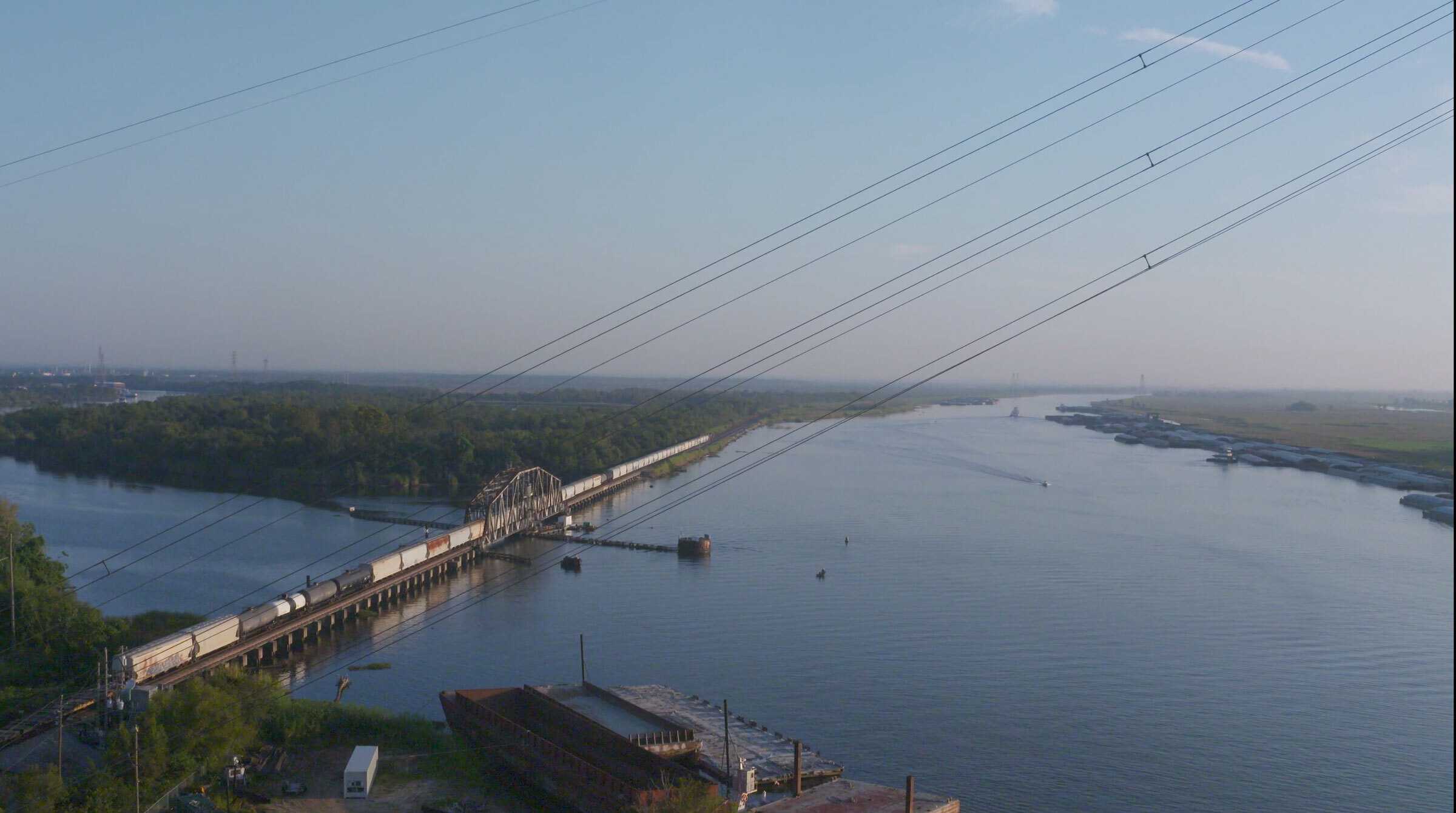 A wide river bordered by forest and wetlands, with a freight train crossing a long bridge. Power lines stretch diagonally across the sky, and barges are moored along the far riverbank.