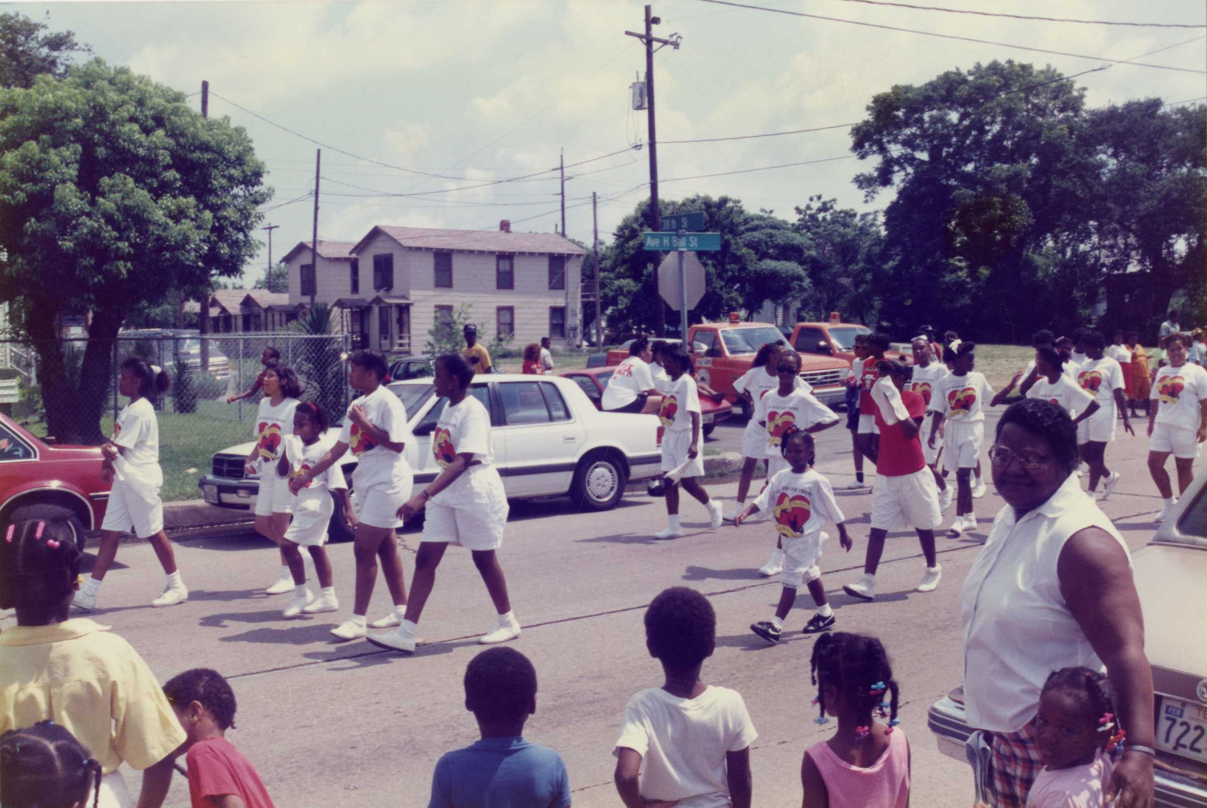Parade with people wearing matching shirts and shorts walking down a street with cars, trees, and onlookers in the background.