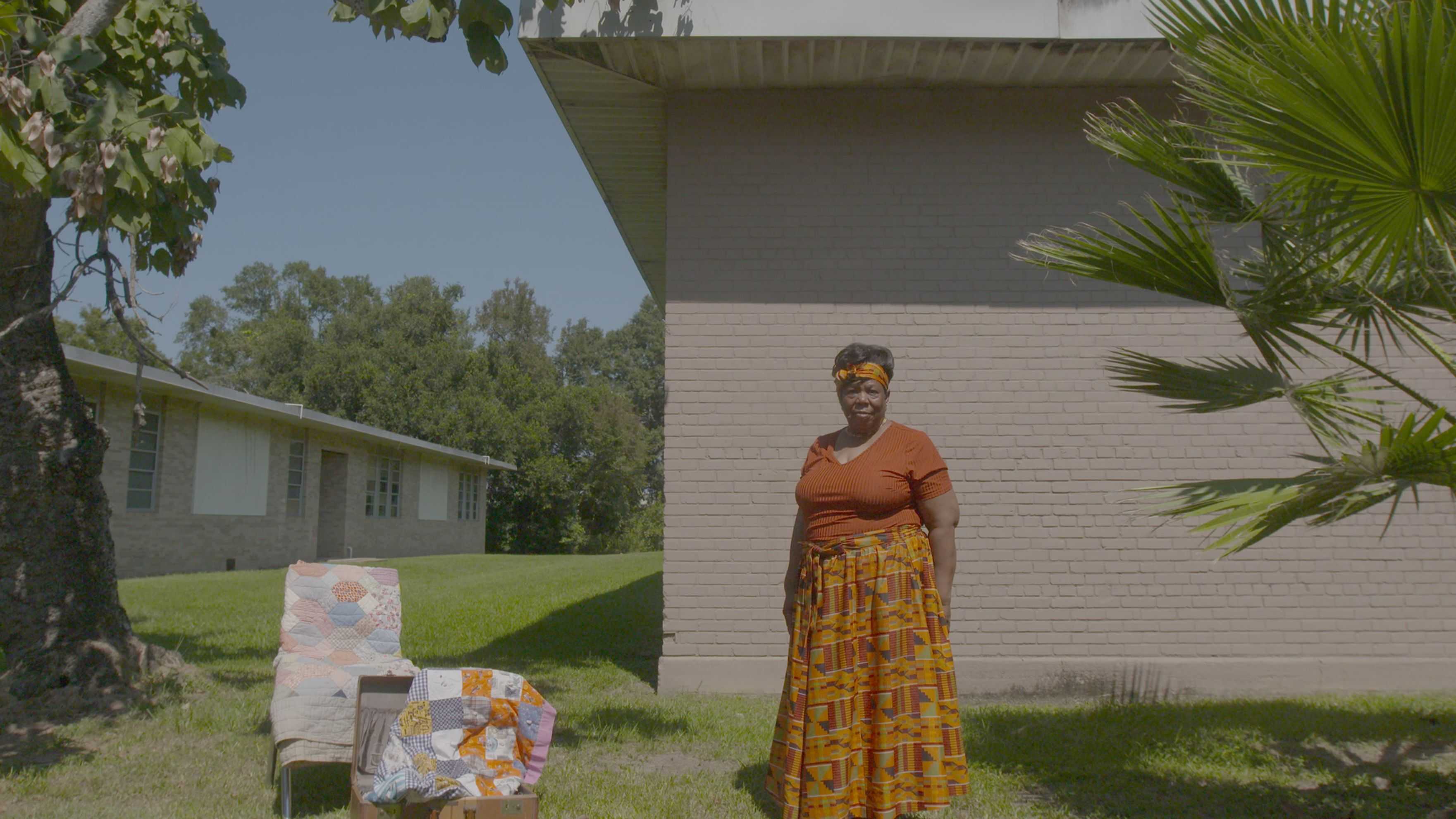 Person standing outdoors beside handmade quilts displayed on a chair and table in front of a building.