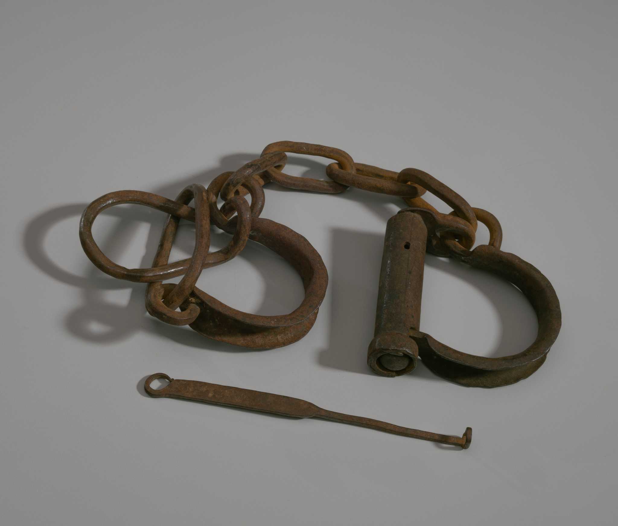 Metal shackle connected by a four link chain to a pair of metal loops. Shackle has a small hole for a key. Key (.2) consists of as thin piece of metal with small loop at end the faces forward. A flat loop is found at the opposite end of the key.