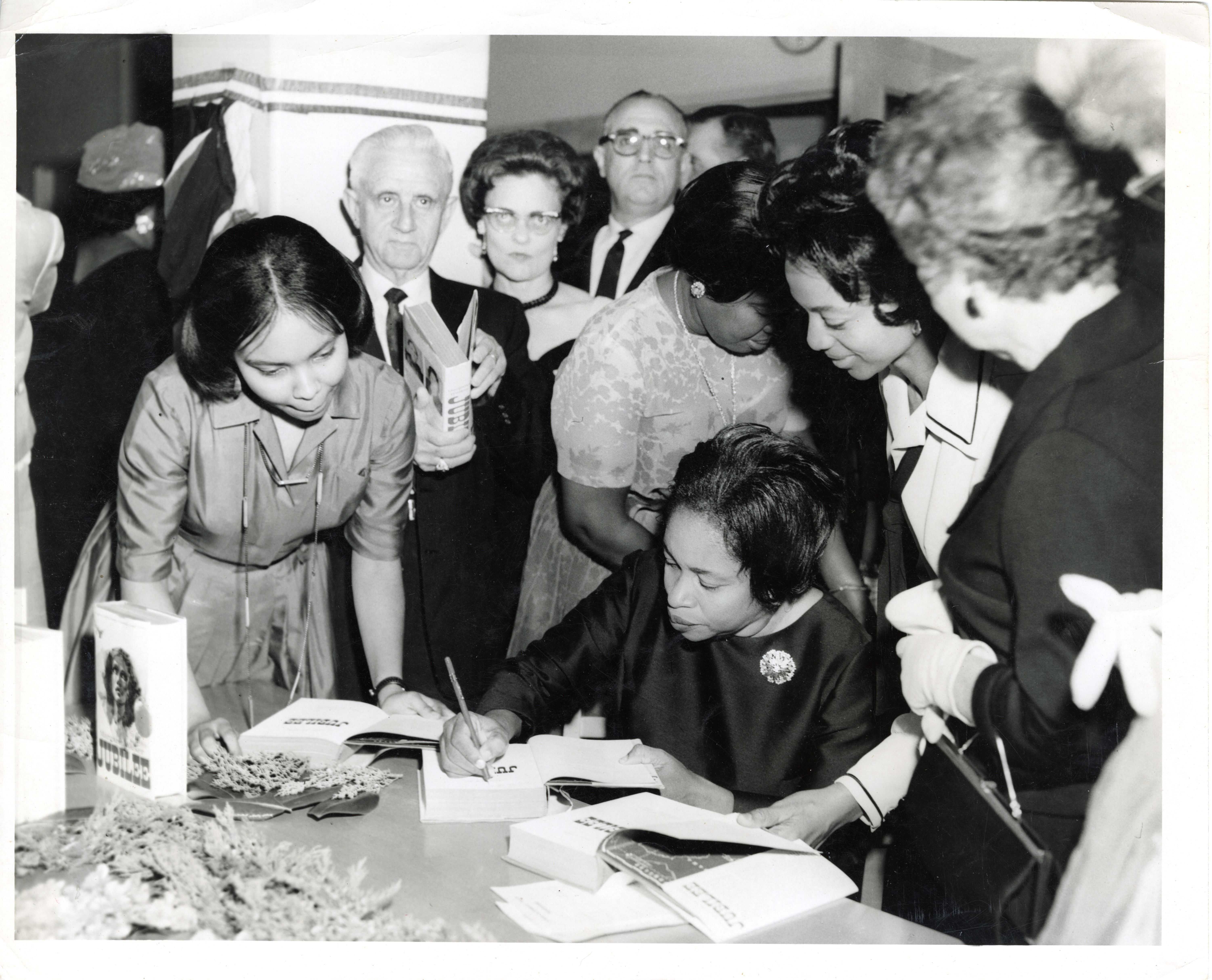 Black and white photo of a book signing event, with a seated individual  (Margaret Walker) signing books at a table surrounded by people holding or observing copies of the same book.