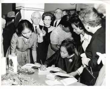 Black and white photo of a book signing event, with a seated individual  (Margaret Walker) signing books at a table surrounded by people holding or observing copies of the same book.