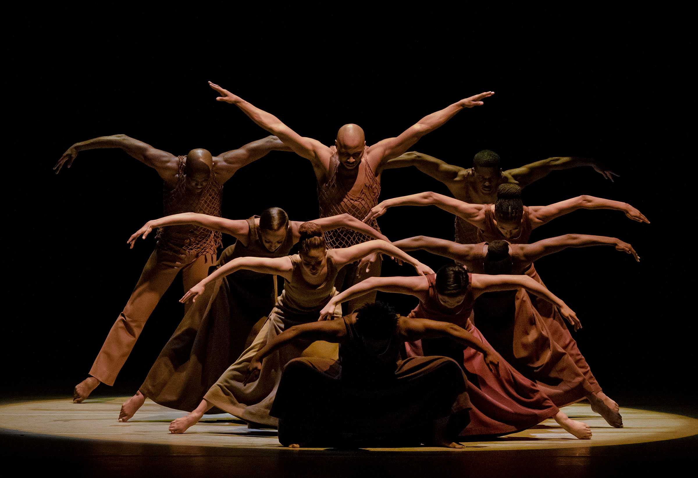 Dancers in a dynamic formation with arms extended, performing on a dimly lit stage.