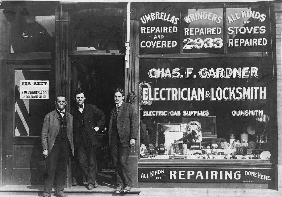 Three men dressed in suits standing before the Chas. F. Gardner Electrician & Locksmith storefront.  The store has writing all over the windows advertising "Umbrellas Repaired and Covered", "Wringers Repaired,"  "All kinds of Stoves Repaired" & "All Kinds of Repairing Done Here"