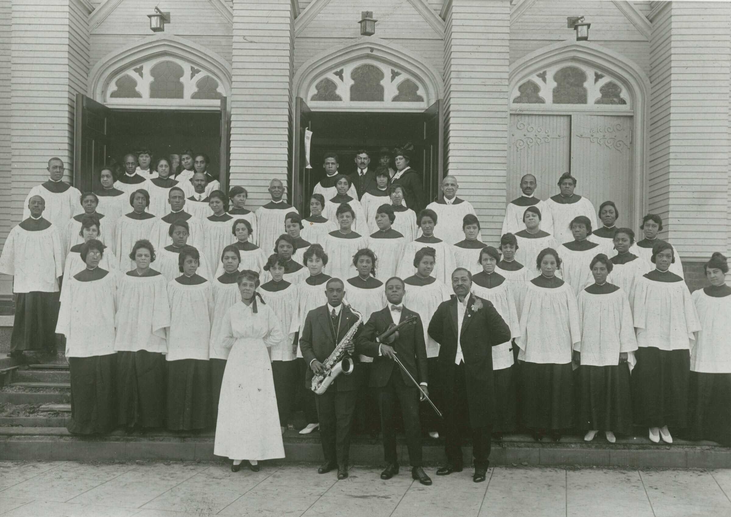 Black and white photograph of a church choir standing in rows on the front steps of a church. The church façade features three (3) doors, the left two (2) are open with people standing in the doorways. Above the doors are gothic style tympanums filled with stained glass windows. The front row of people standing on the sidewalk are three (3) men and one (1) woman. The woman stands on the far PR and wears a white dress. Around her neck is a bow positioned over her shoulder. The three (3) men wear dark suits with white shirts. The two (2) men in the middle hold instruments. The PR musician holds a saxophone and the PL musician holds a violin. The man on the far PL stands with his hands held behind his back. On the reverse in the lower left corner is a penned note [First AME Church/Rev. J. Logan Craw, pastor, and Choir/COLLECTION OF MIRIAM MATTHEWS]. In the upper right corner is the number [4] horizontally oriented.