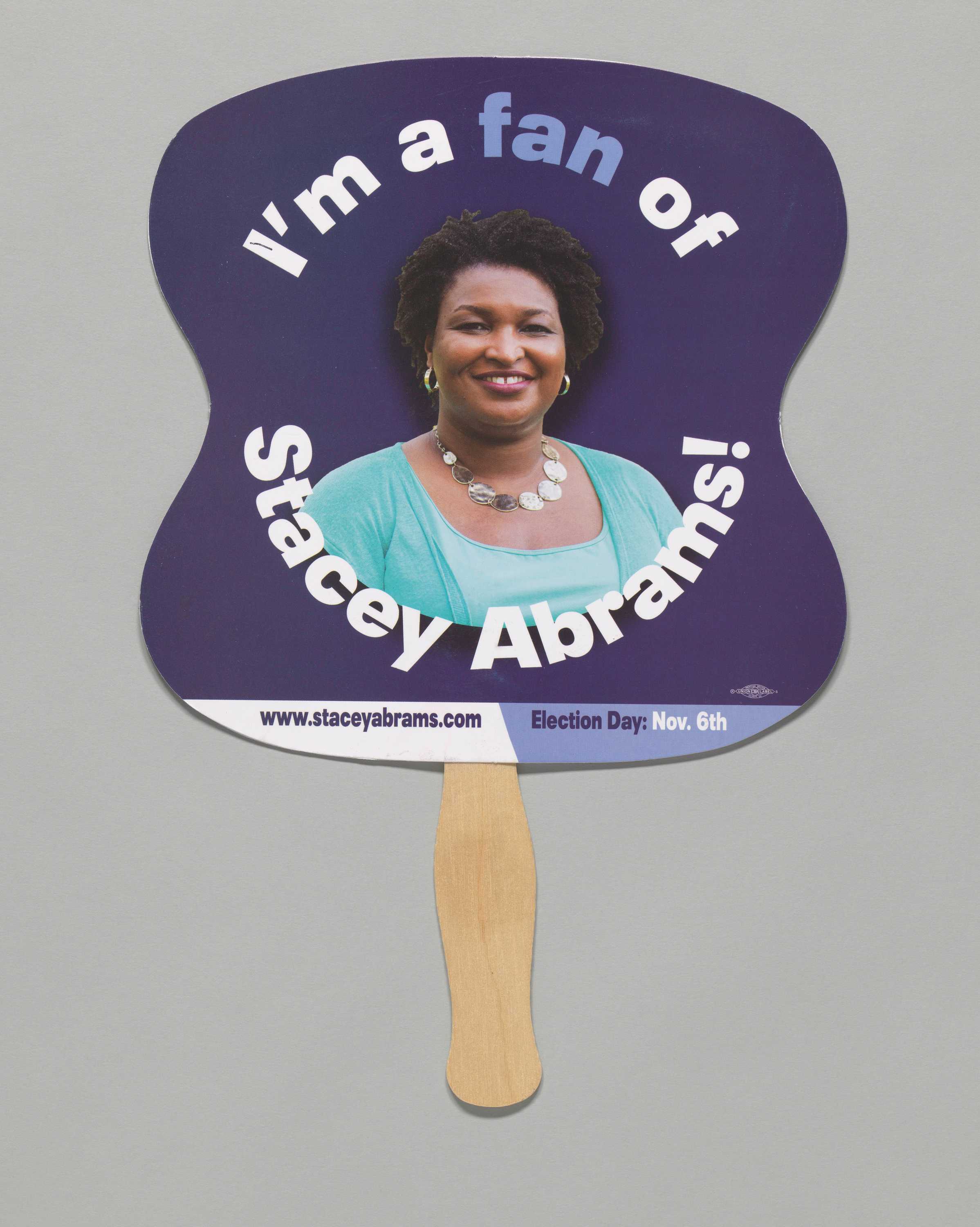 A dark blue paper fan from the Stacey Abrams 2018 Georgia gubernatorial campaign, mounted on a flat wooden handle.