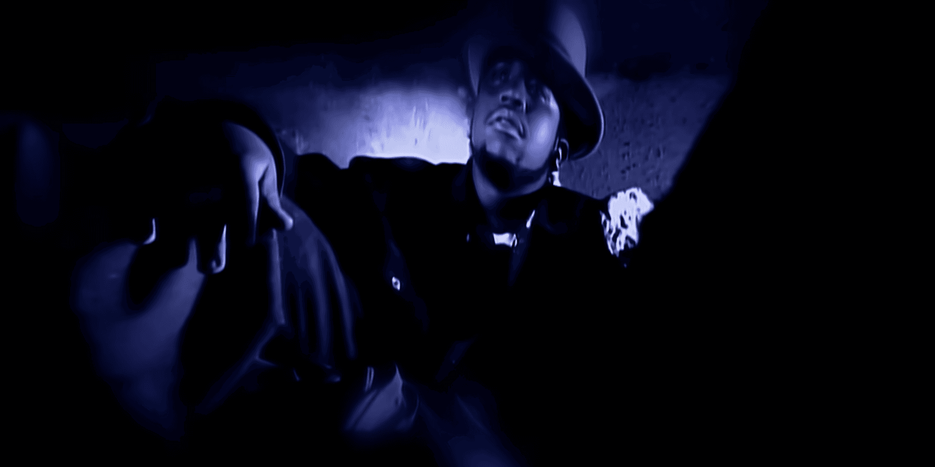Big Boi is wearing a bowling hat while sitting wide leg on the floor against blue hue lighting.