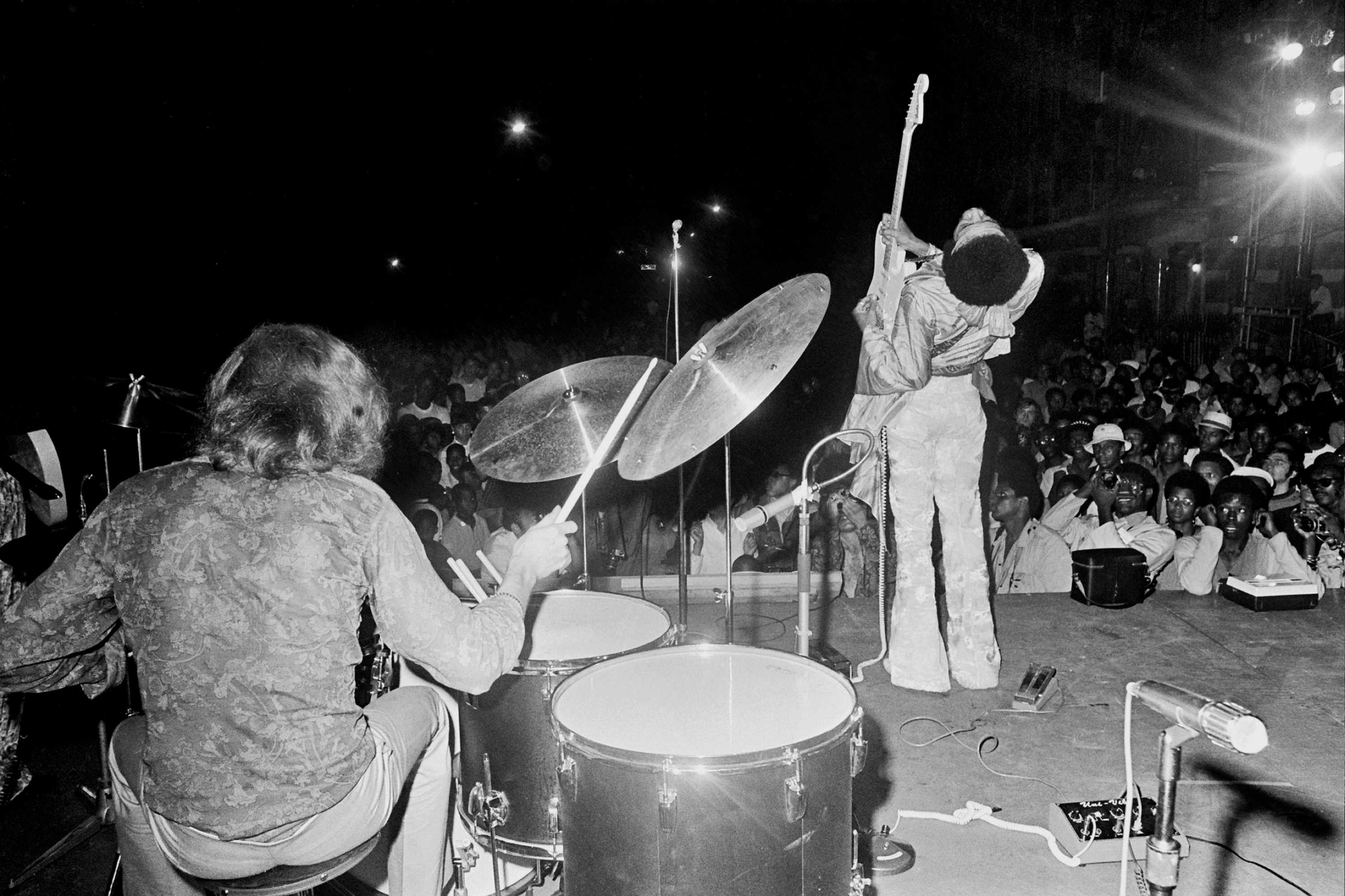 A photograph from the back of the stage as Hendrix is playing to an audience. His drummer is in the left corner of the frame.