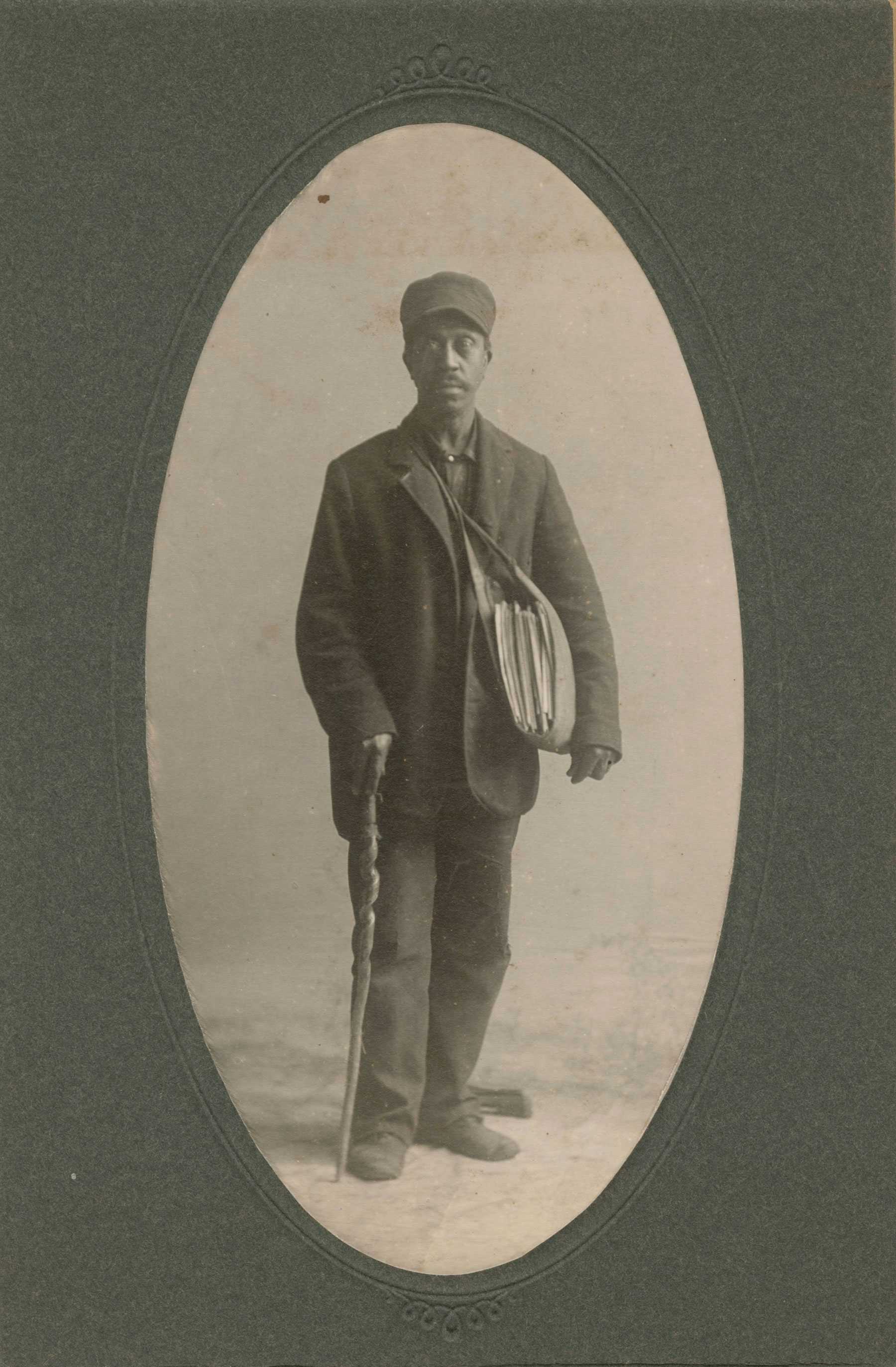 Sepia-toned portrait of a man in a cap and coat, holding a walking stick, with a satchel over his shoulder.