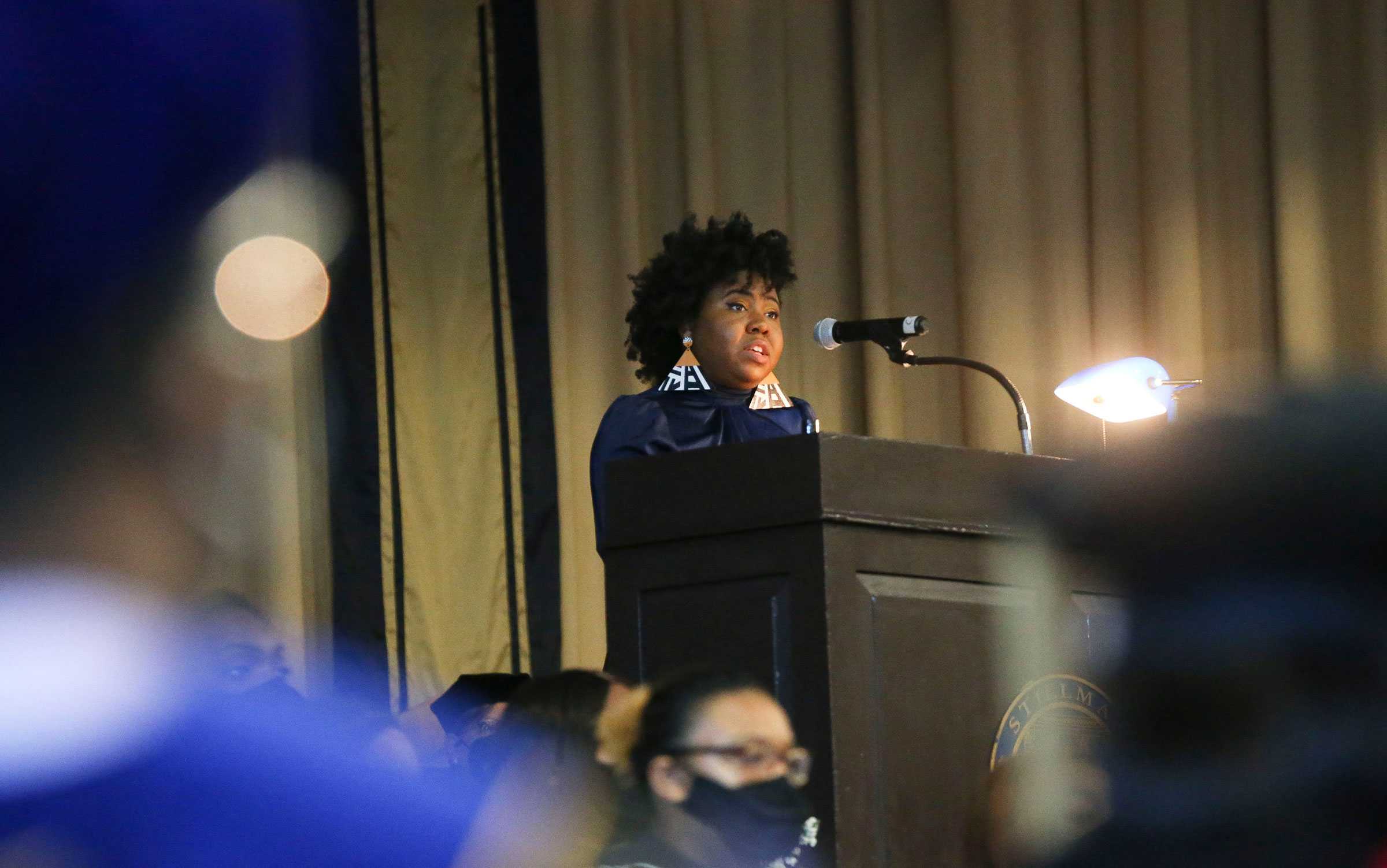 Person speaking at a podium with a microphone, in an indoor setting with audience members visible.