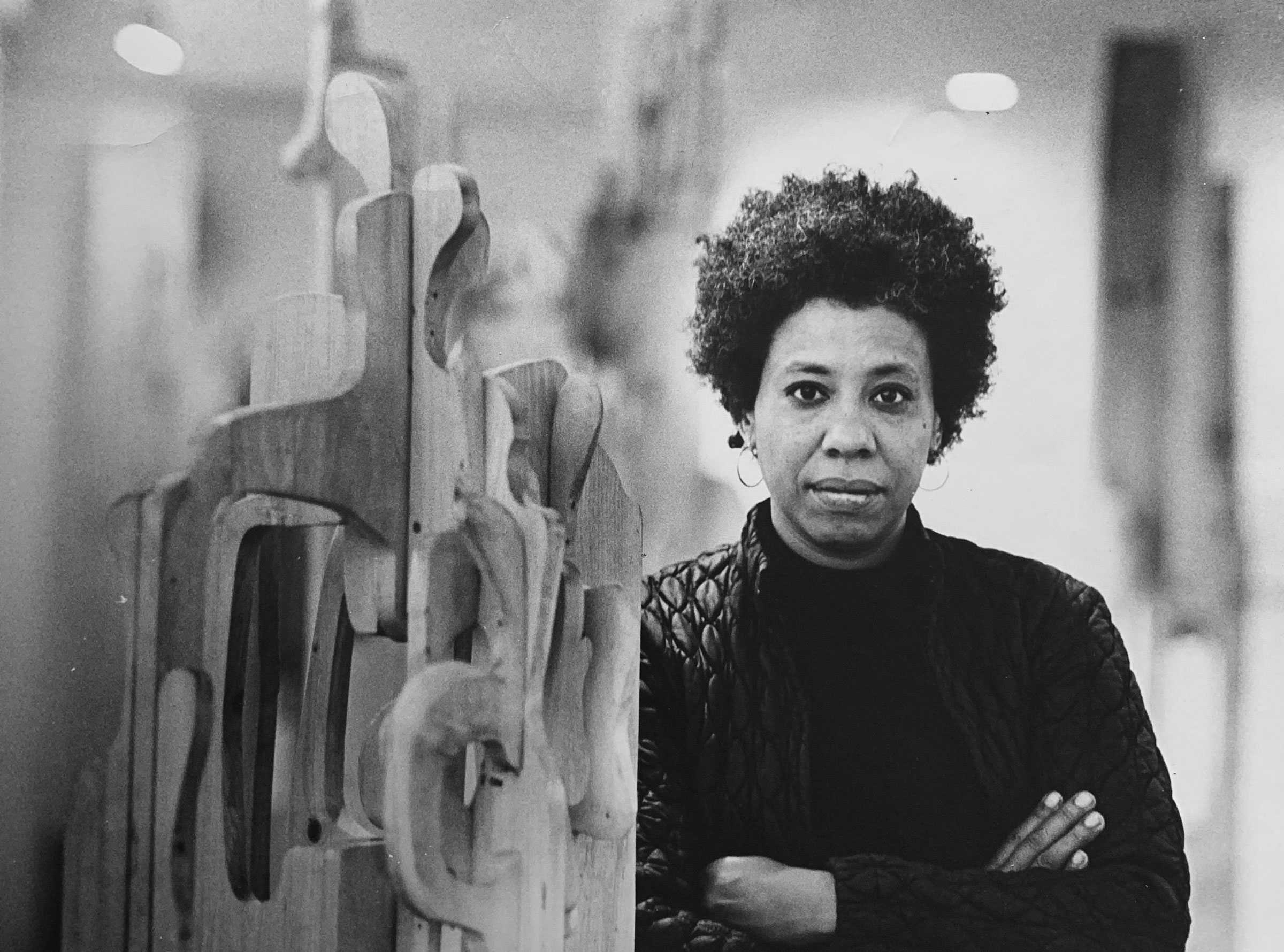 Black-and-white photo of a woman with short curly hair, standing beside a wooden sculpture.