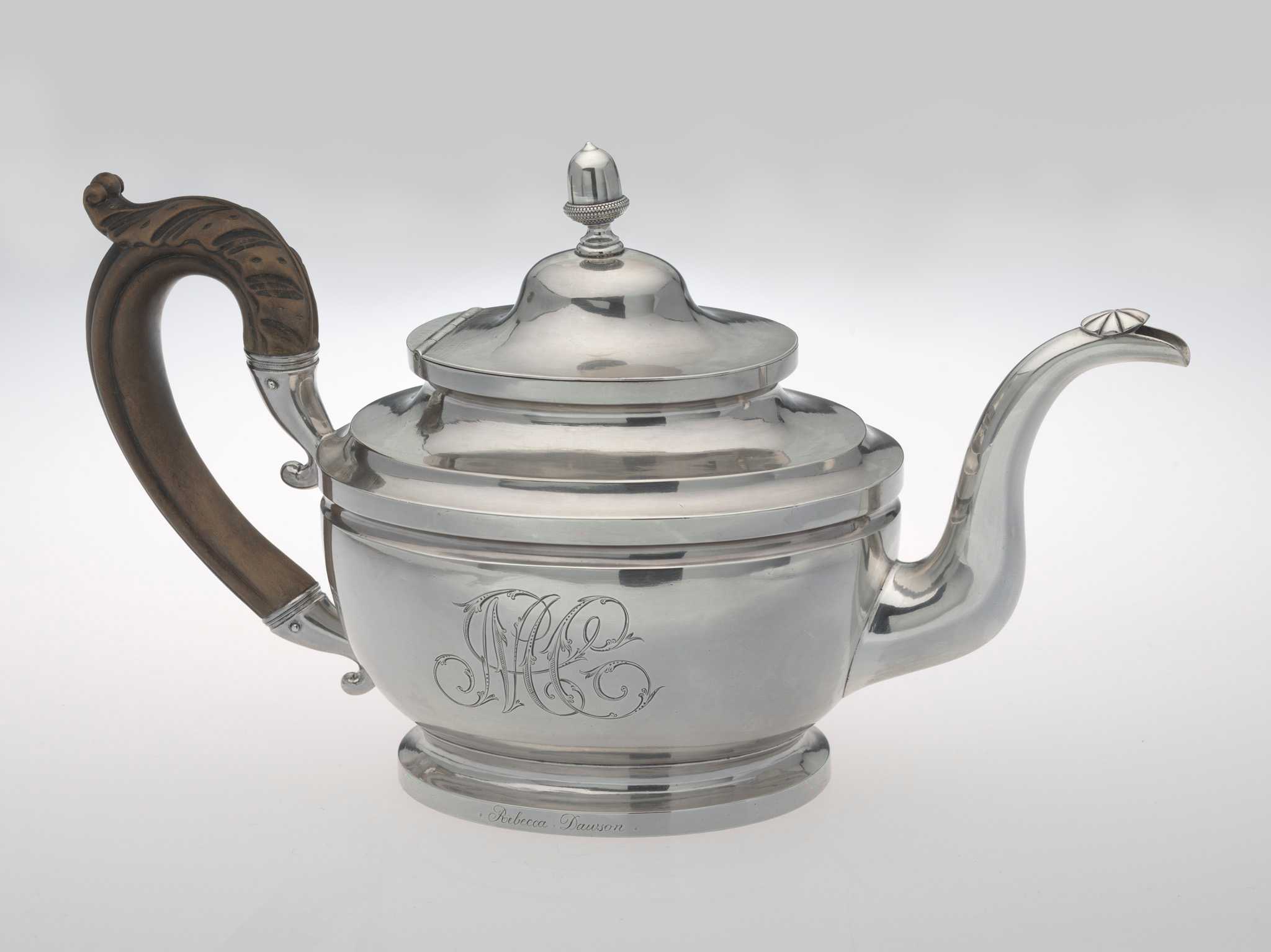 The silver teapot has an oval vase-shape on a spreading pedestal foot, with curved spout capped by an incised patera and wooden leaf-capped scroll handle, and hinged domed cover with acorn finial. The scripted monogram "MC" has been engraved on the side and the name "Rebecca Dawson" has been engraved along the bottom rim.