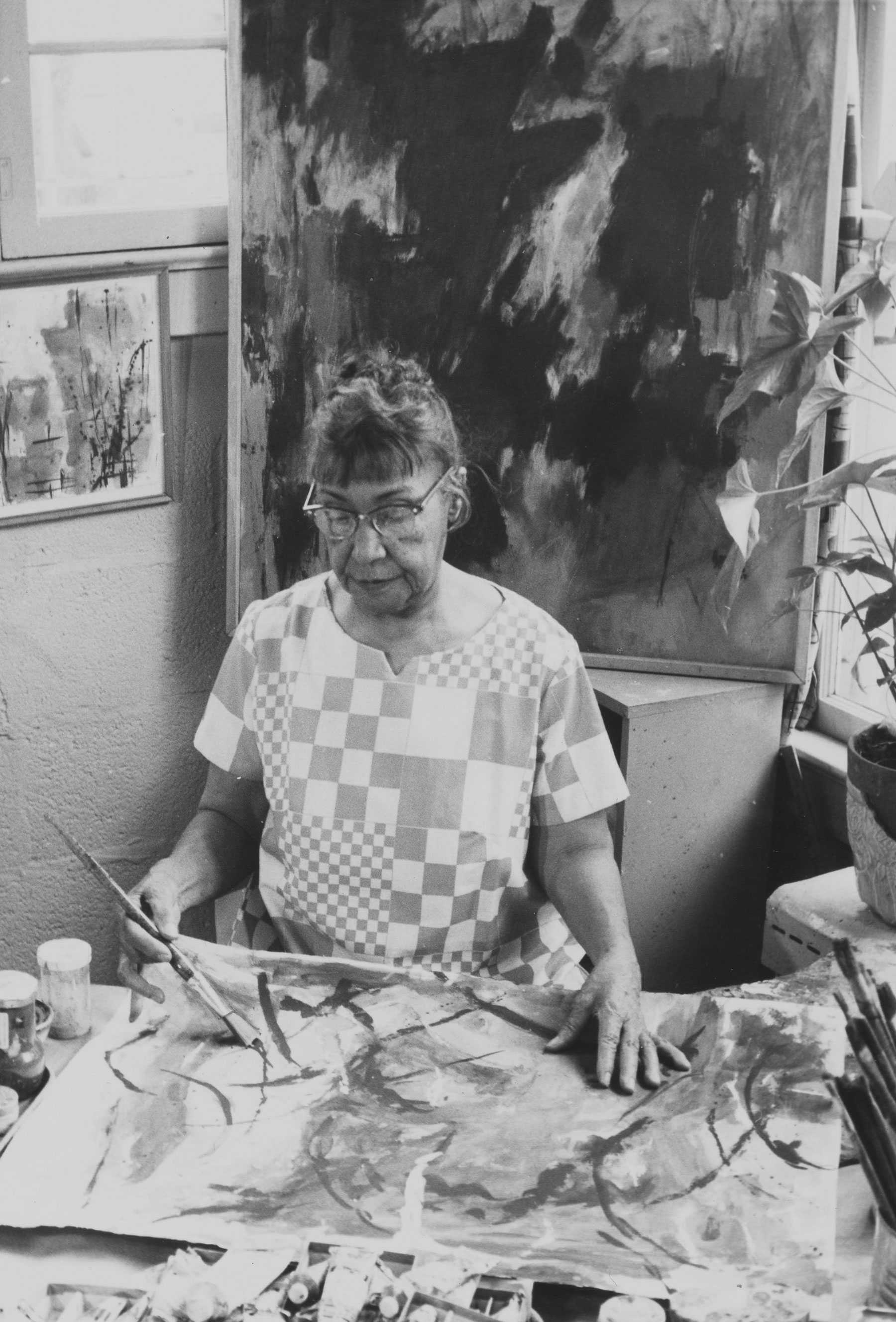 Black-and-white photo of a woman painting at an easel in her art studio.