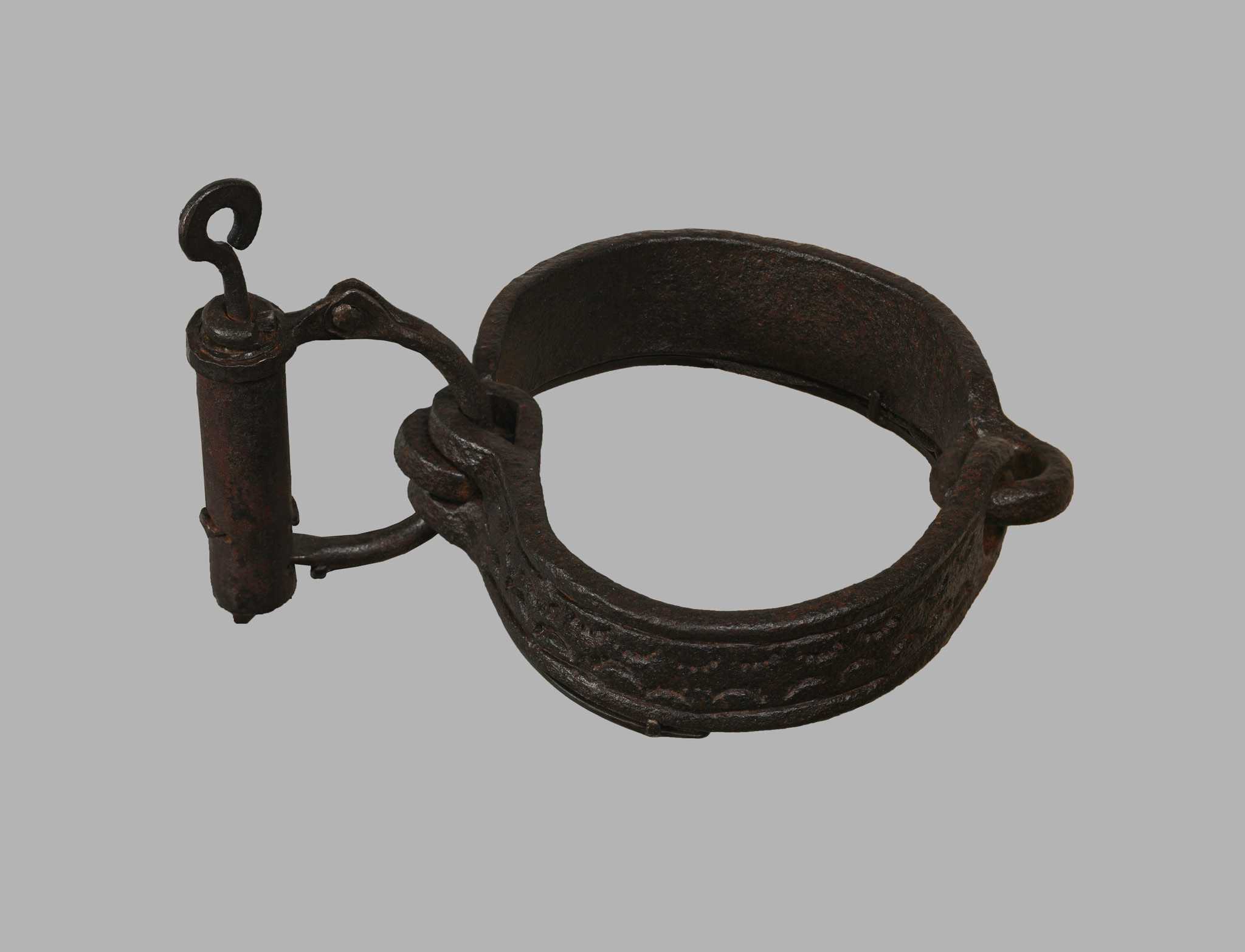 This wrought iron collar is formed of two pieces of semicircular iron joined together by two interlocking metal loops. At each end of the two pieces are another two rings. Through these are looped a D-shaped ring attached to a locking mechanism. The outer surface of the two pieces has been incised with a repeating semi-circular design.