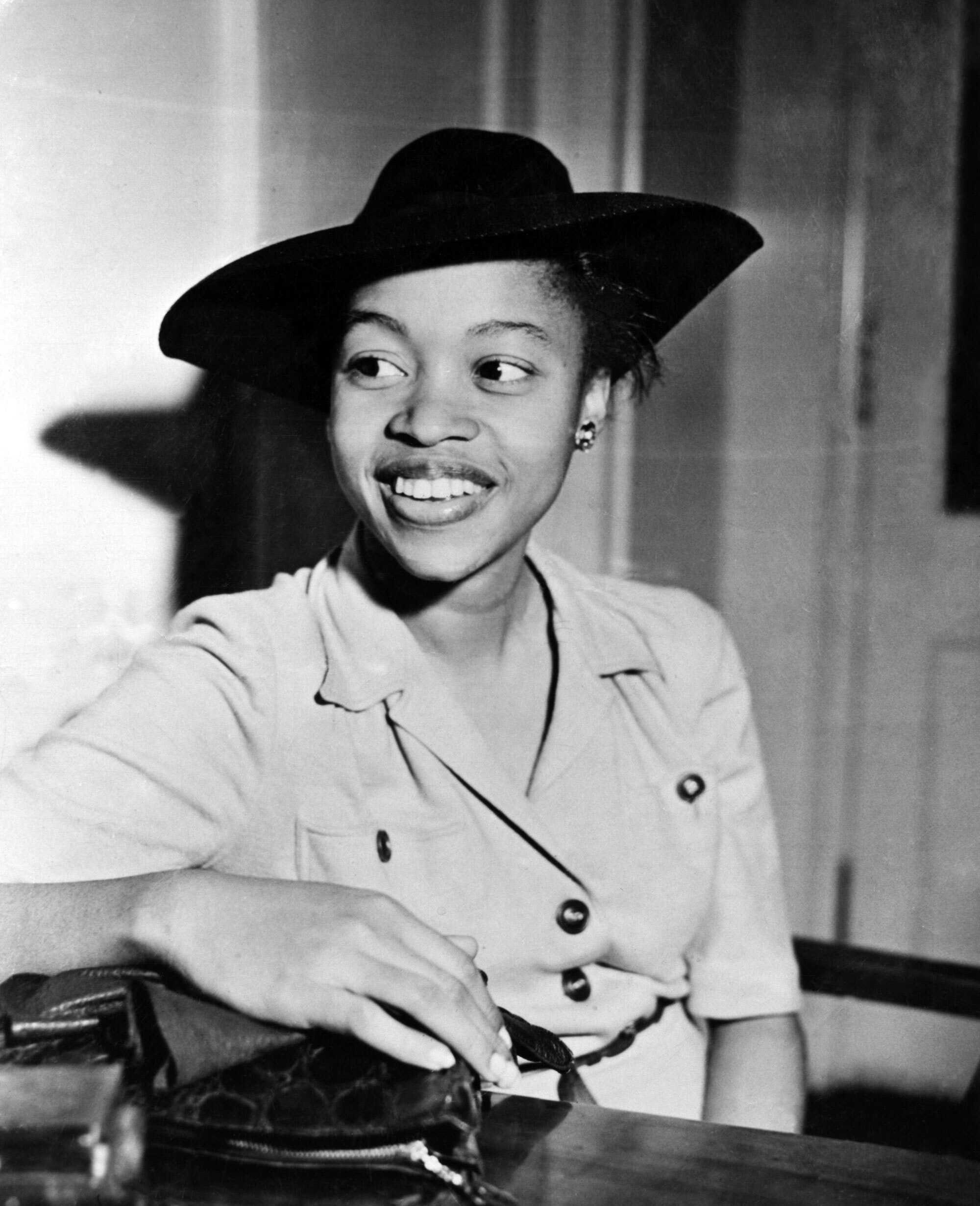 Author Margaret Walker seated at a table wearing a light-colored buttoned dress and a dark wide-brimmed hat, looking to the side with an open-mouthed expression.