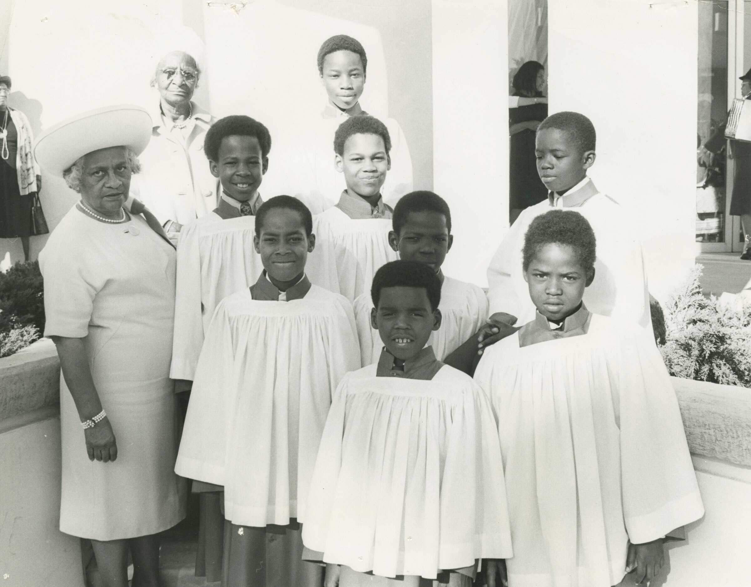 Black and white photograph of a group of young boys dressed in altar clothes. The group includes eight (8) boys in several different rows. The boys wear white robes and dark skirts and dark collars over their clothing. On the left side of the image are two (2) older women wearing white looking at the camera, one in the foreground and one in the background. the group stands outside with the church interior seen in the upper right quadrant. On the reverse in the lower right corner on the bottom in pen [ALTAR BOYS].
