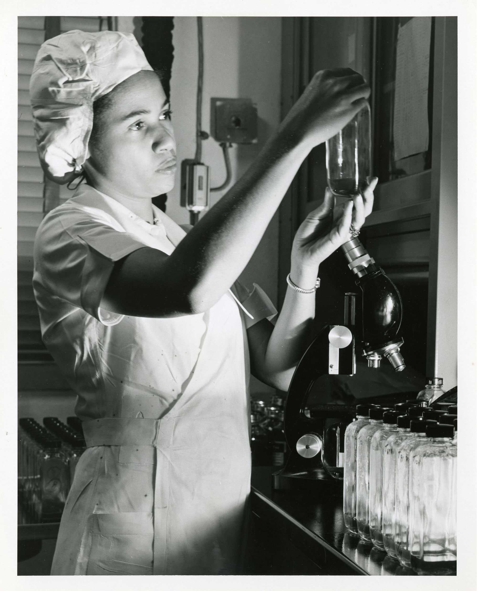 Photograph of a lab technician examining a glass bottle near a microscope, symbolizing scientific research.