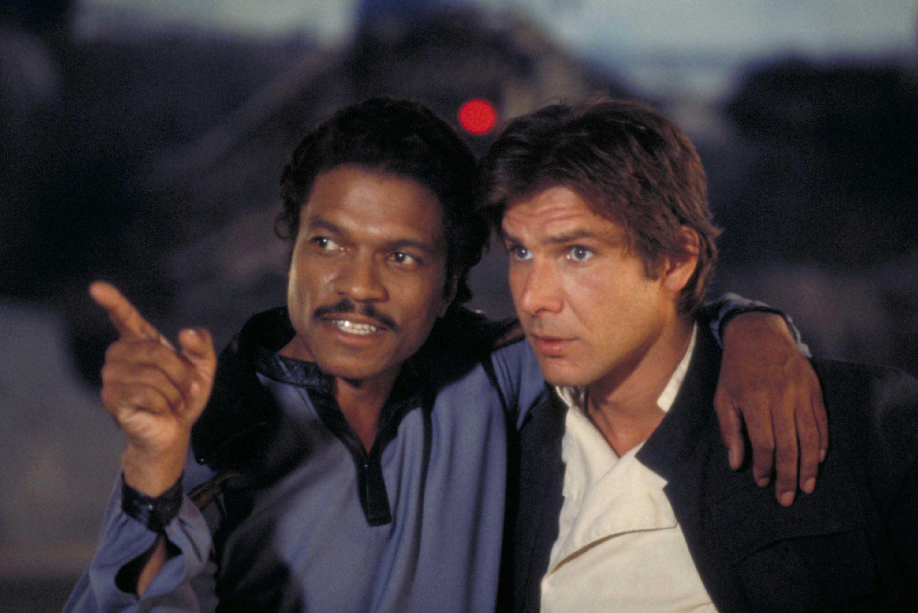 Billy Dee Williams has his arm around Harrison Ford pointing to something off in the distance.