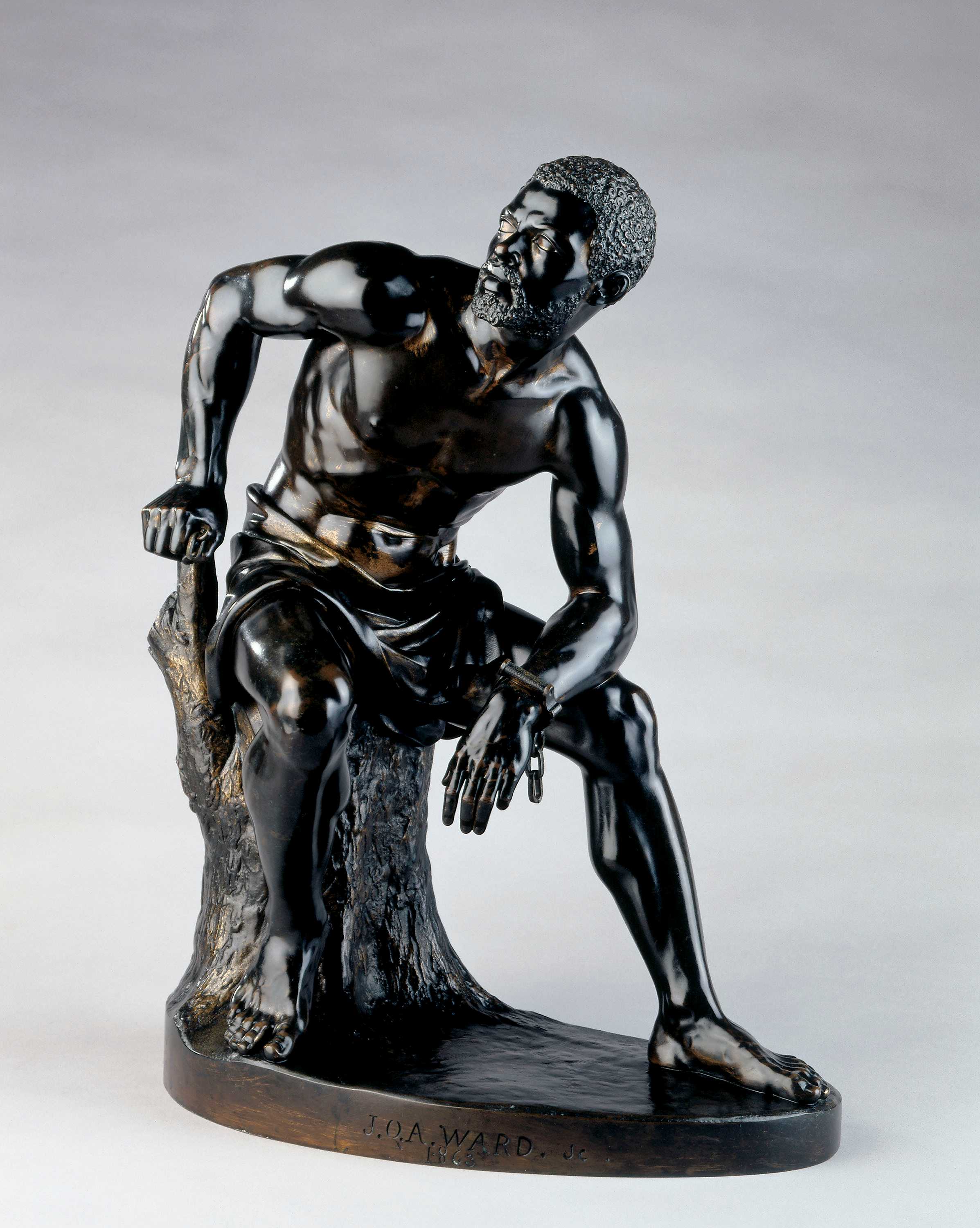 A bronze statue of an shirtless African American man sitting on a stump. On his left wrist is a single handcuff, the chain dangling down.