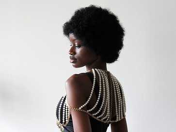 Person in profile wearing a black garment adorned with draped strands of white pearls connected by gold chains across the shoulders and back.