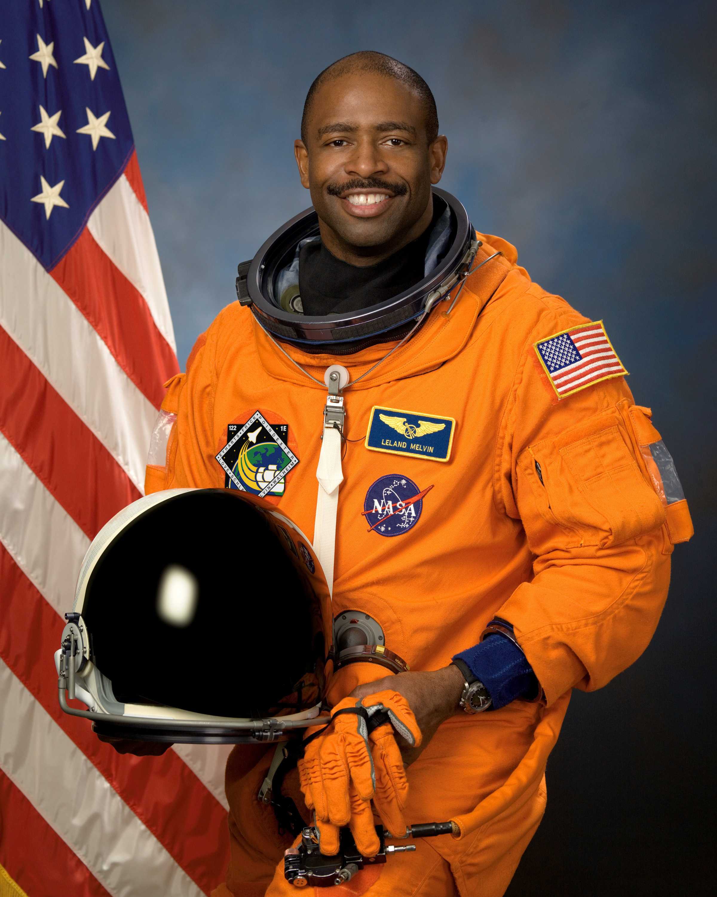 Melvin posed for his portrait while wearing an orange space suit and a helmet and gloves in his hands.