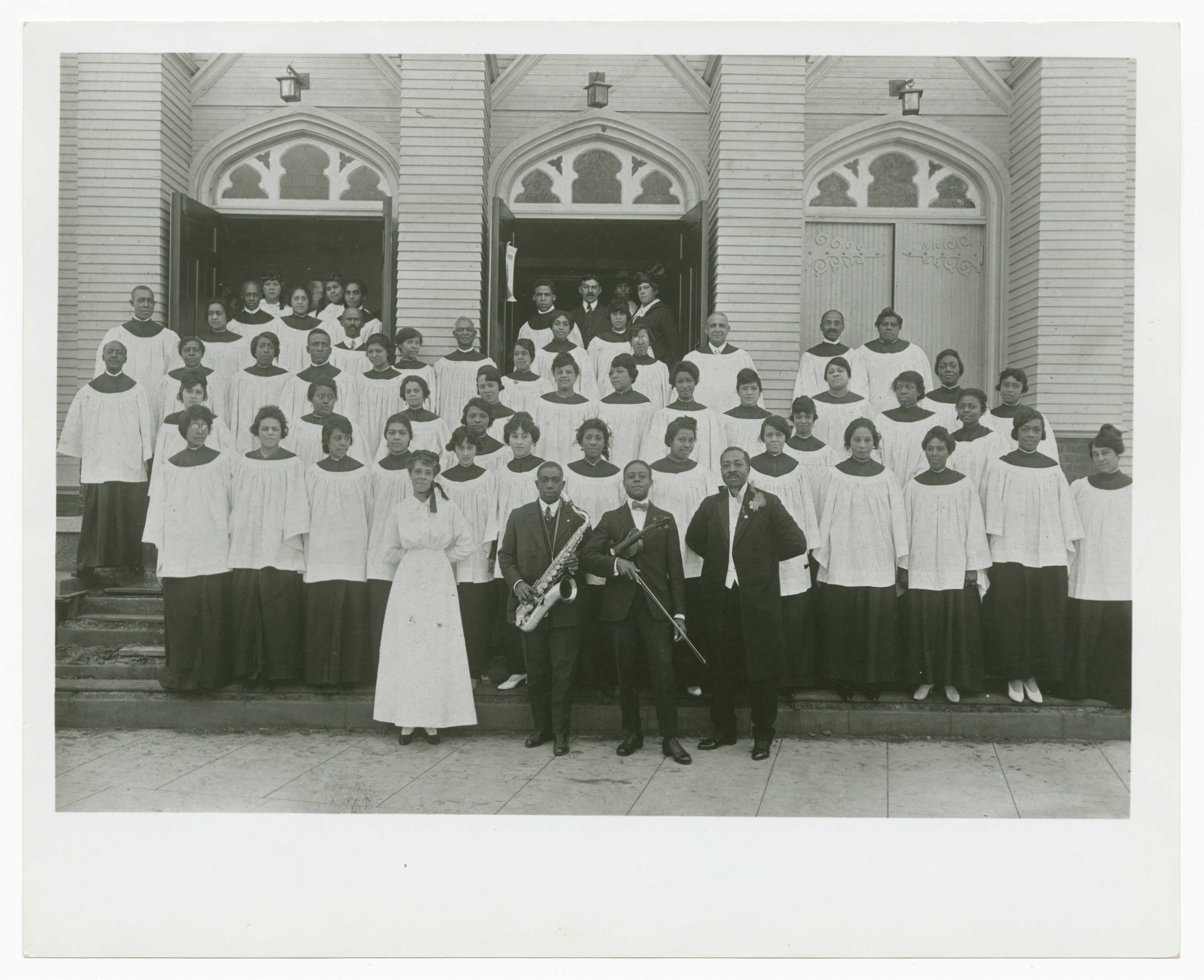 Black and white photograph of a church choir standing in rows on the front steps of a church. The church façade features three (3) doors, the left two (2) are open with people standing in the doorways. Above the doors are gothic style tympanums filled with stained glass windows. The front row of people standing on the sidewalk are three (3) men and one (1) woman. The woman stands on the far PR and wears a white dress. Around her neck is a bow positioned over her shoulder. The three (3) men wear dark suits with white shirts. The two (2) men in the middle hold instruments. The PR musician holds a saxophone and the PL musician holds a violin. The man on the far PL stands with his hands held behind his back. On the reverse in the lower left corner is a penned note [First AME Church/Rev. J. Logan Craw, pastor, and Choir/COLLECTION OF MIRIAM MATTHEWS]. In the upper right corner is the number [4] horizontally oriented.